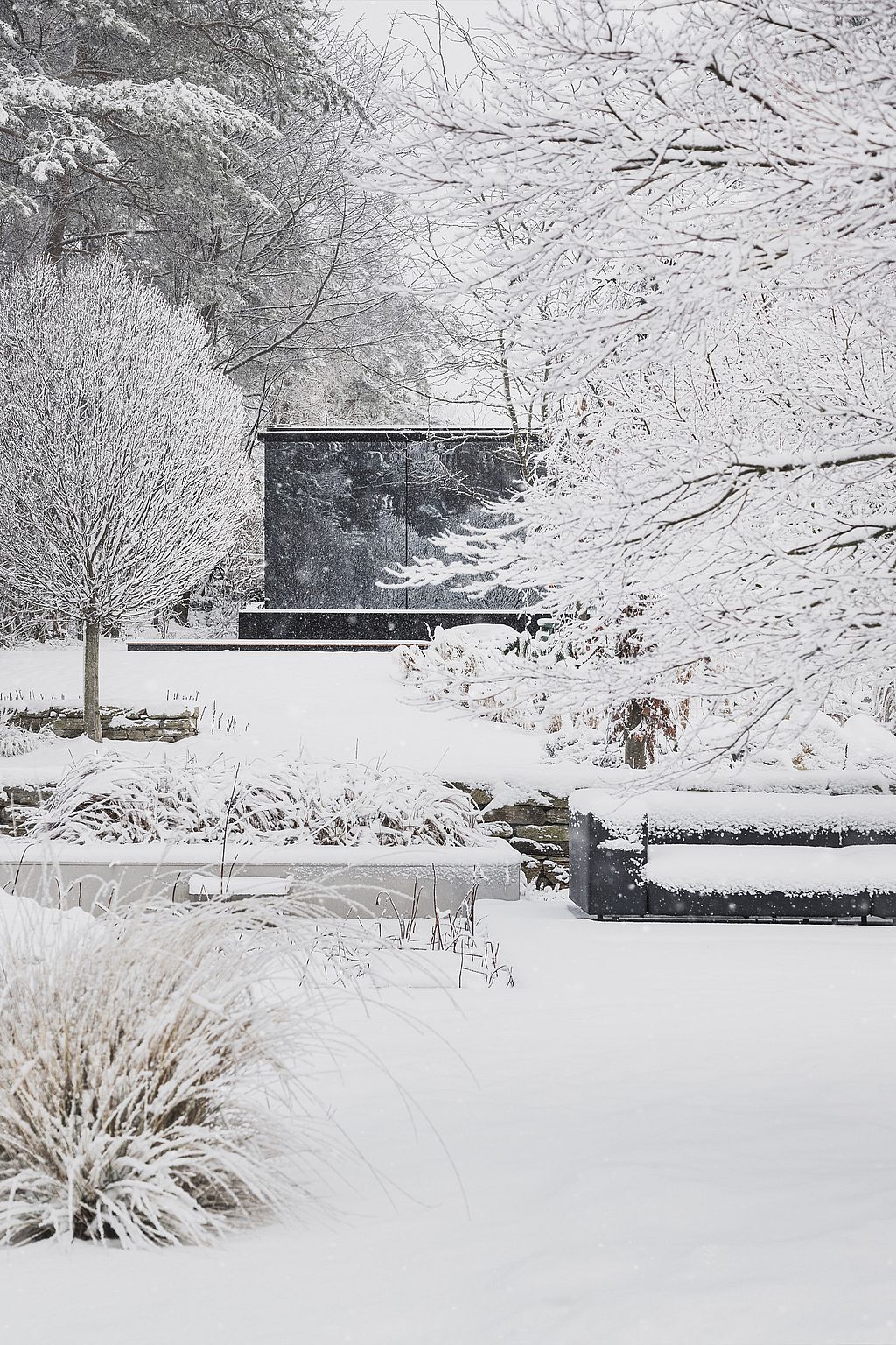 Verschneiter Wintergarten mit dunklem Außensofa unter frostbedeckten Bäumen, eingerahmt von Ziergräsern und geschichteten Pflanzbeeten.