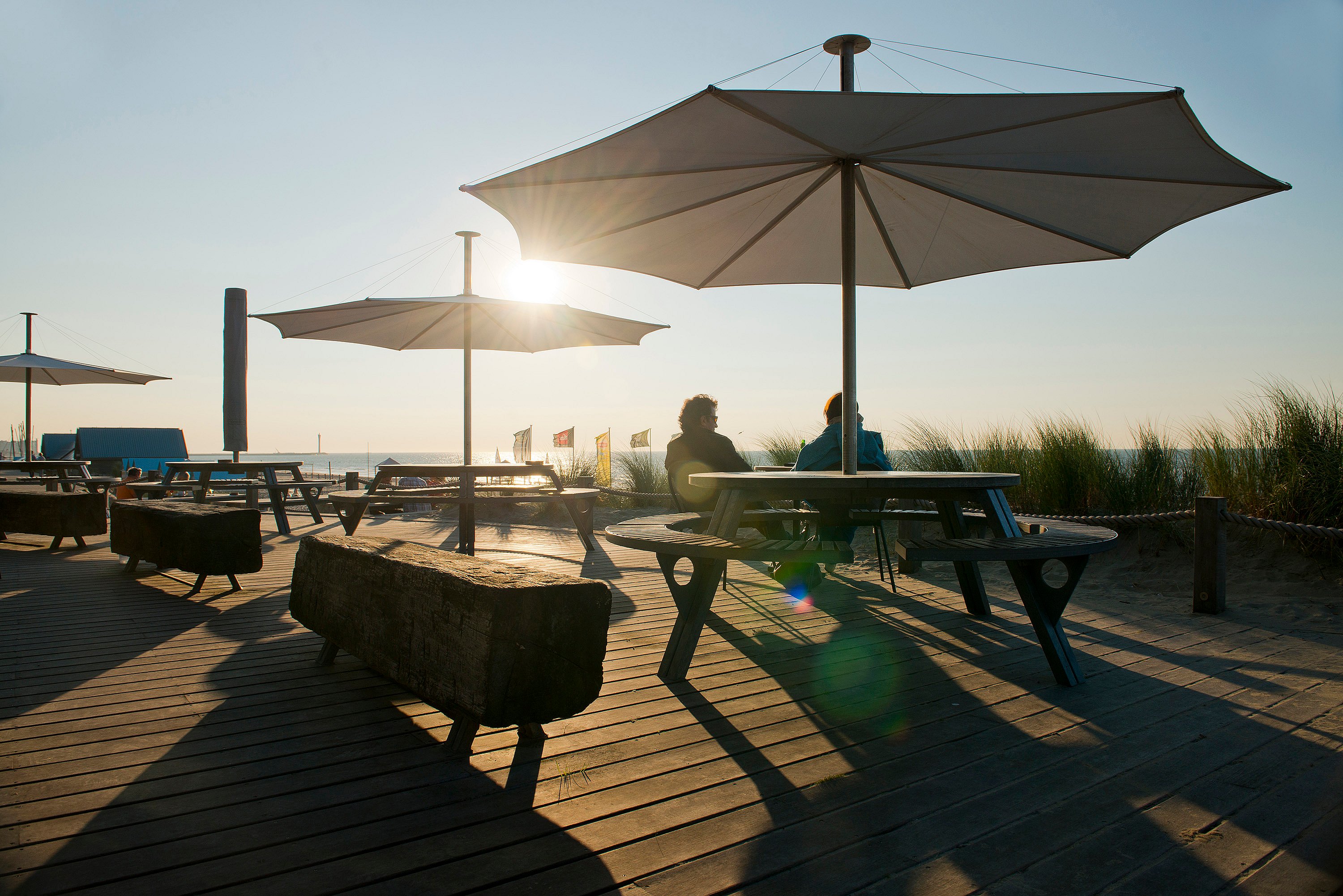 Bij zonsondergang staan ​​twee Gargantua picknicktafels op een strandterras onder grote parasols. Aan één tafel zitten twee mensen en lange schaduwen strekken zich uit over de houten planken.
