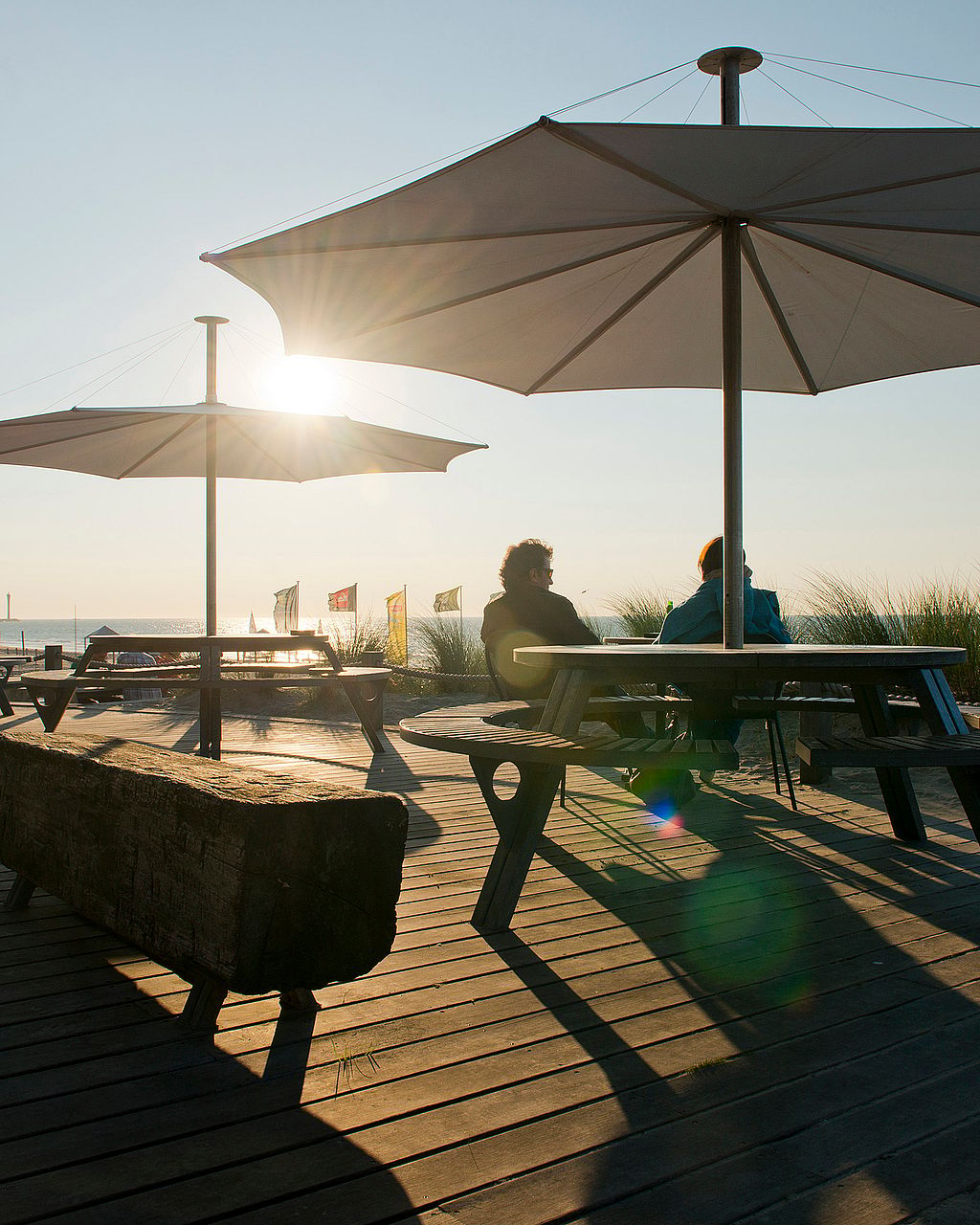 Au coucher du soleil, deux tables de pique-nique Gargantua sont installées sur une terrasse de plage, sous de grands parasols. Deux personnes sont assises à l'une d'elles, et de longues ombres s'étirent sur les planches de bois.