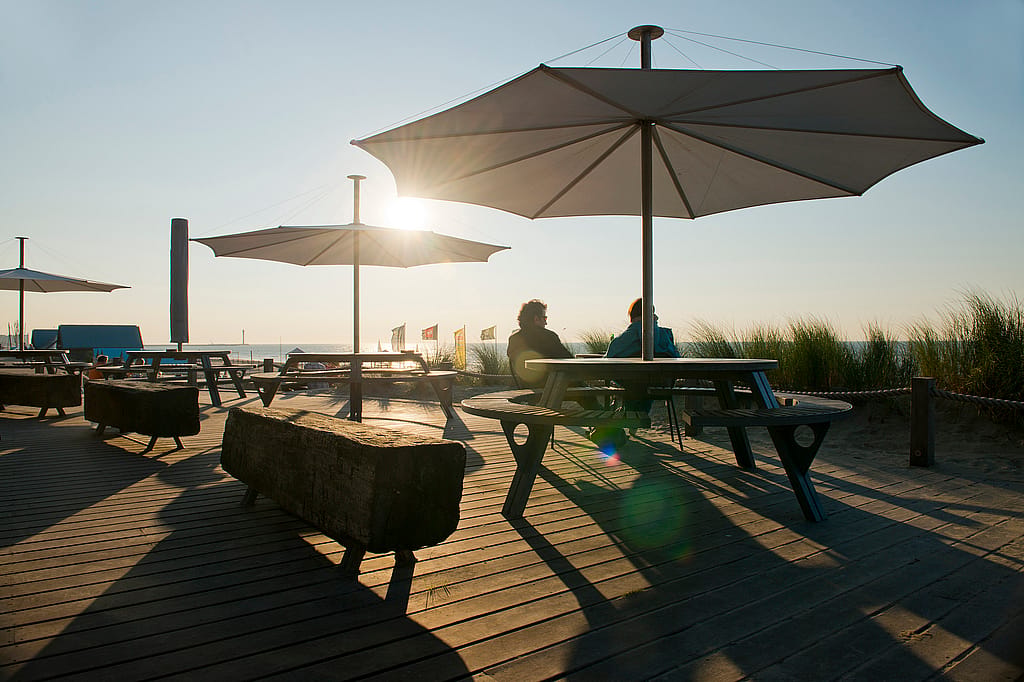 Au coucher du soleil, deux tables de pique-nique Gargantua sont installées sur une terrasse de plage, sous de grands parasols. Deux personnes sont assises à l'une d'elles, et de longues ombres s'étirent sur les planches de bois.