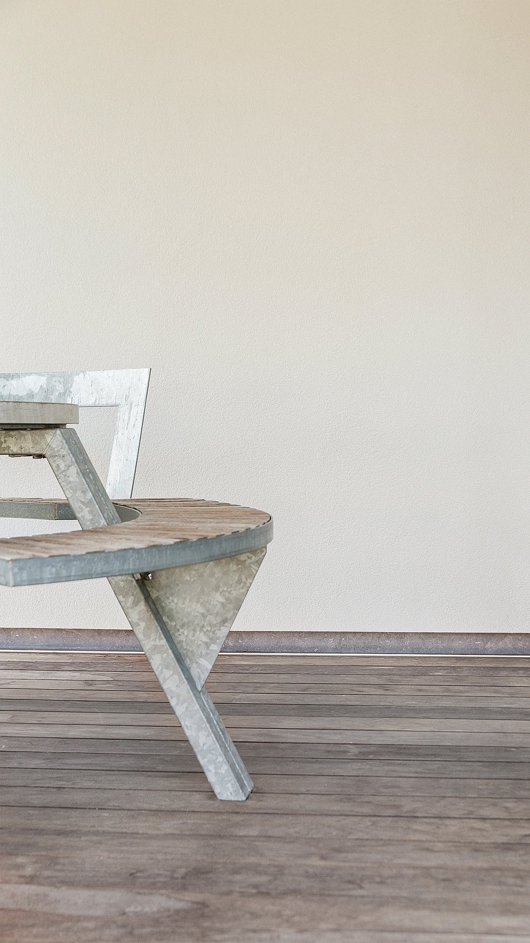   Close-up van de verweerde houten bank en de gegalvaniseerde poot van een Gargantua tafel op een houten terras tegen een effen muur.