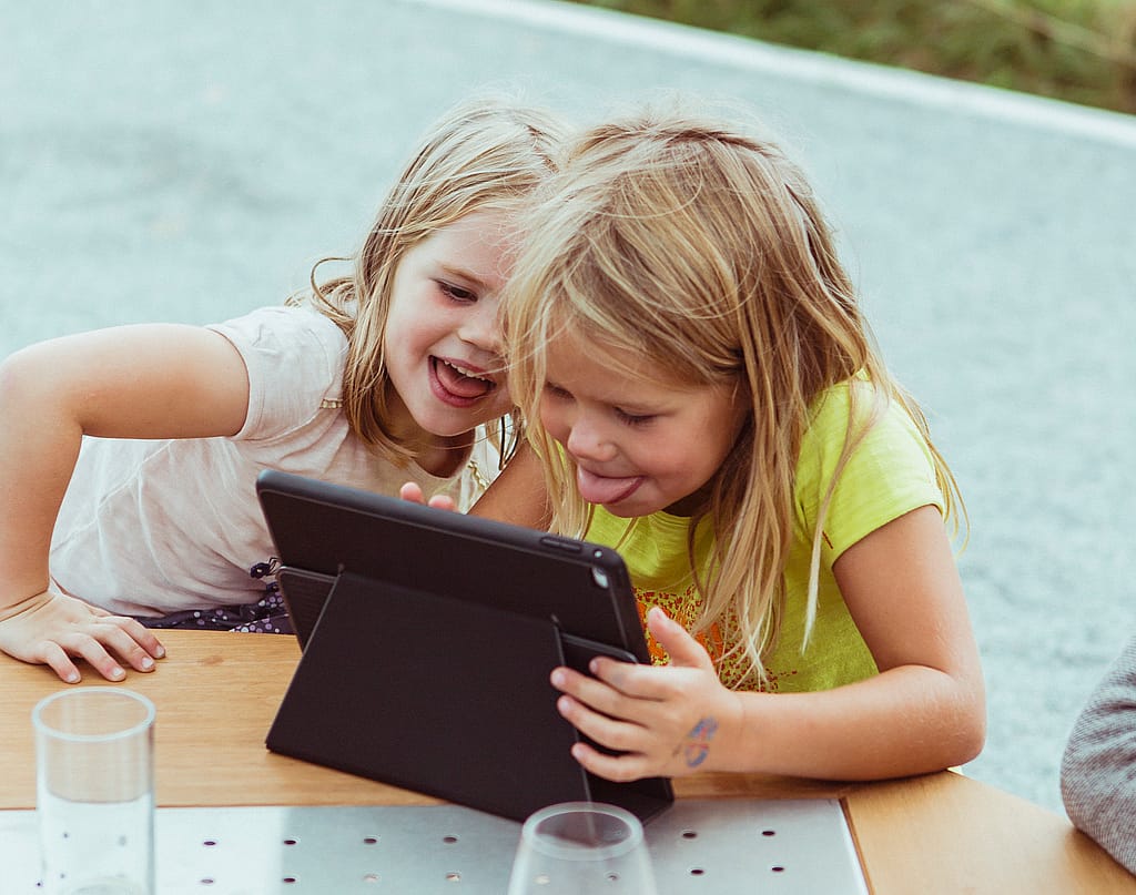 Deux jeunes filles se penchent sur une tablette posée sur le plateau en métal perforé et le bord en bois d'une table Gargantua. Des verres sont posés sur le plateau au premier plan.