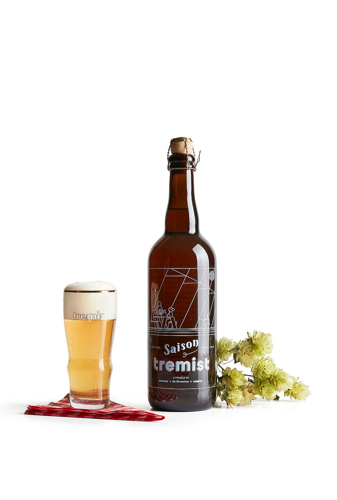 Stillleben aus dem Studio: Eine Flasche und ein Glas Saison Tremist Bier neben hellen Hopfenblüten vor weißem Hintergrund.
