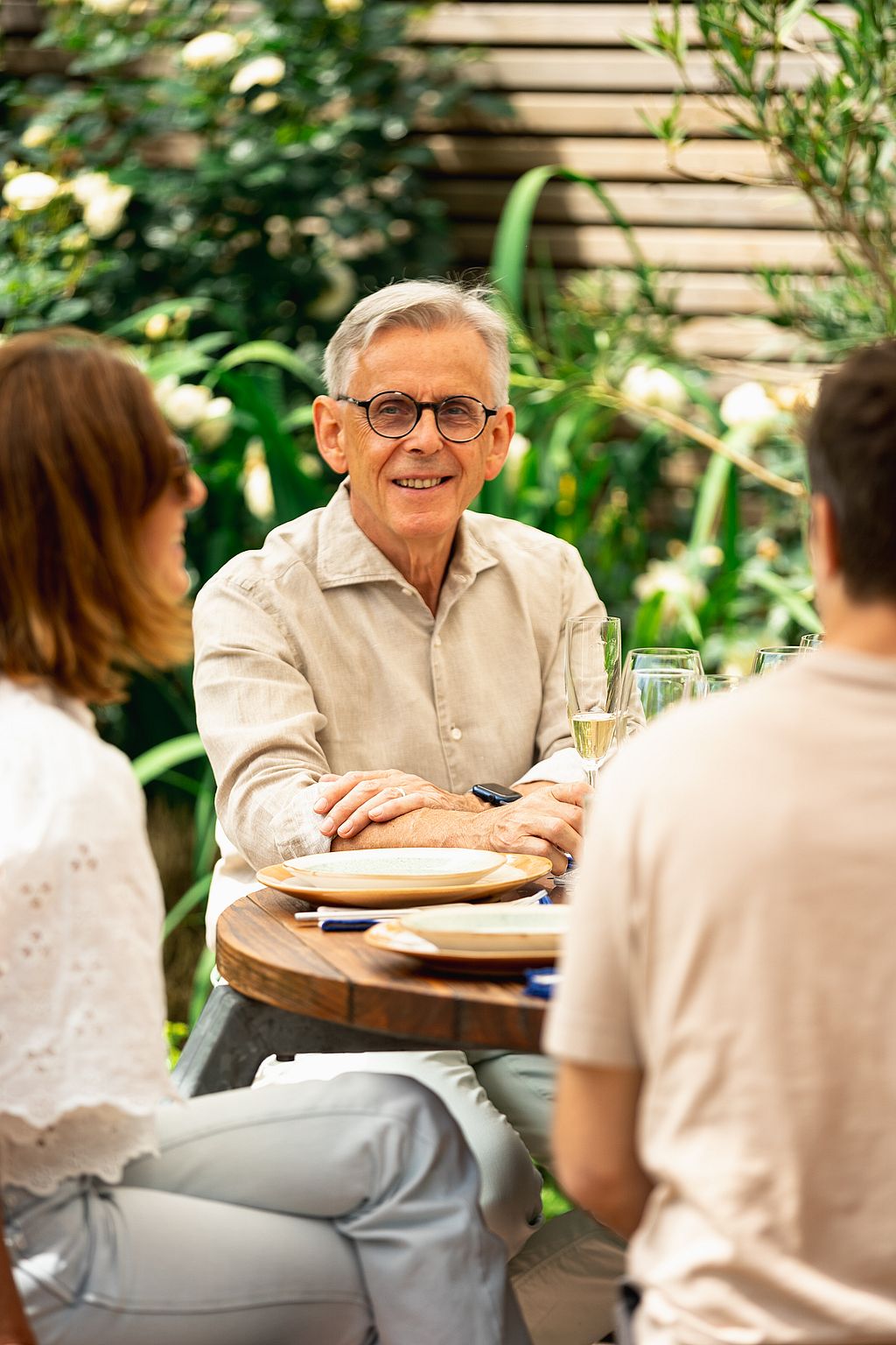 Familieleden zitten rond een Gargantua-tafel in een beplante binnentuin, terwijl een jong meisje naar hen toe loopt.