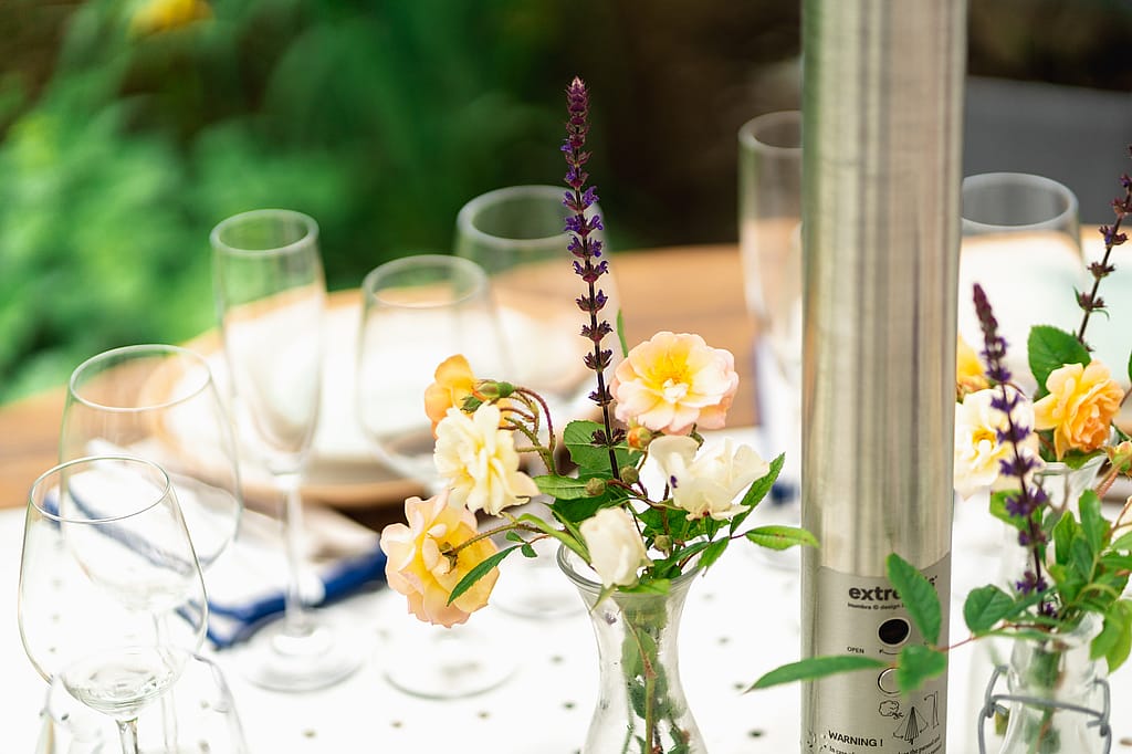 Close-up van bloemen in kleine vazen en lege glazen op het geperforeerde midden van een Gargantua-tafel naast de parasolpaal