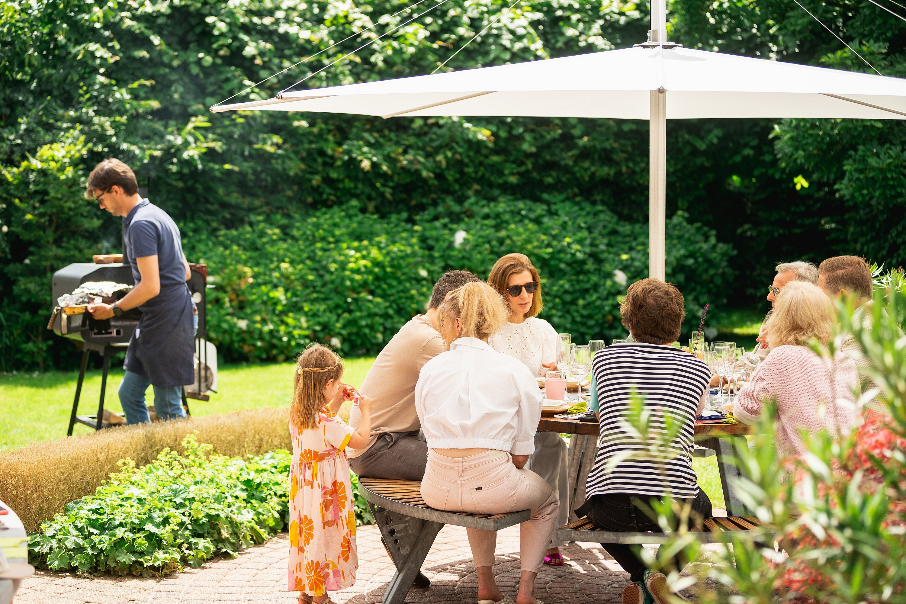 Een gezin verzamelt zich rond een ronde Gargantua-picknicktafel onder een witte Inumbra-parasol in een weelderige tuin, terwijl iemand voedsel in de buurt grilt.
