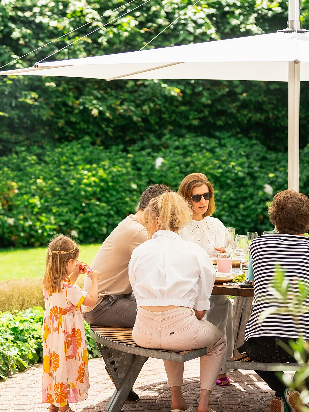 Een gezin verzamelt zich rond een ronde Gargantua-picknicktafel onder een witte Inumbra-parasol in een weelderige tuin, terwijl iemand voedsel in de buurt grilt.