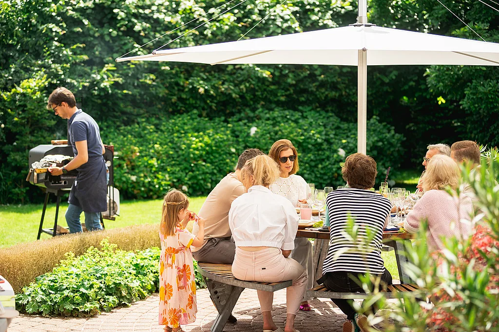 A family gathers around a round Gargantua picnic table under a white Inumbra parasol in a lush garden, while someone grills food nearby.