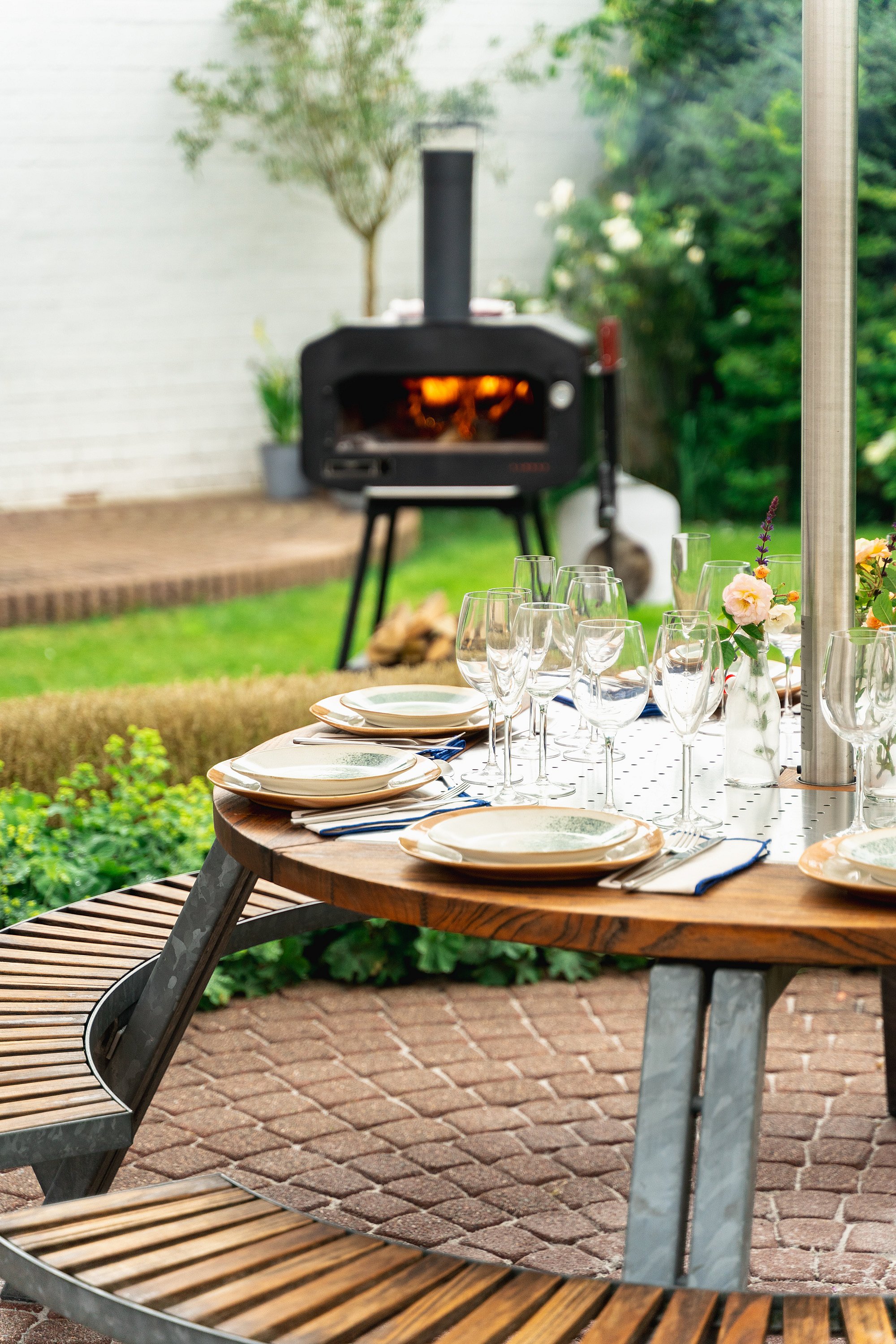 Close-up van een set Gargantua tafel met borden, glazen en bloemen, met een verlichte barbecue zacht wazig op de achtergrond.