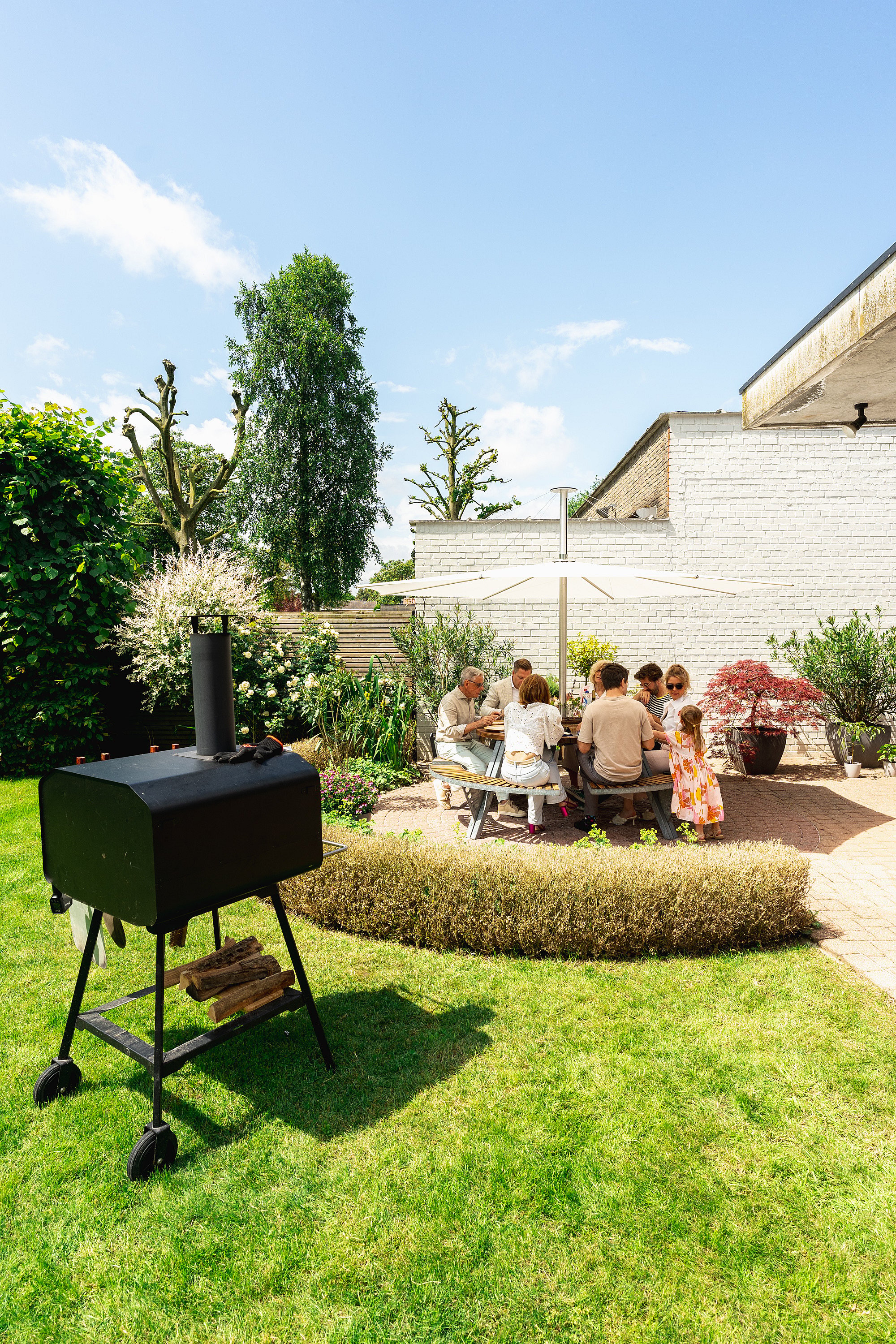 Breed uitzicht op een zonnige tuin met een ronde Gargantua picknicktafel onder een witte Inumbra parasol, omgeven door witte bakstenen muren, plantbedden en een barbecue op de voorgrond.