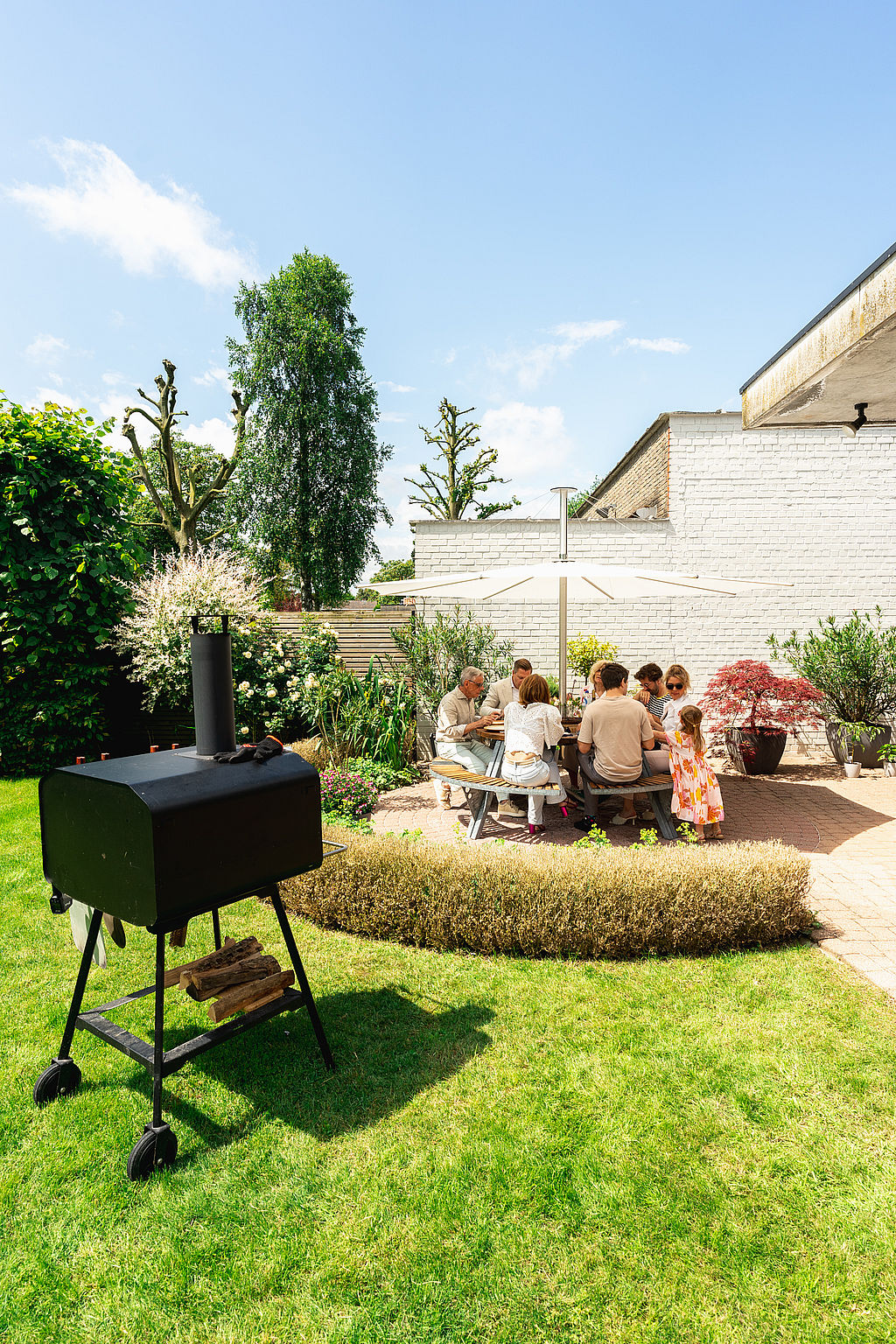 Breed uitzicht op een zonnige tuin met een ronde Gargantua picknicktafel onder een witte Inumbra parasol, omgeven door witte bakstenen muren, plantbedden en een barbecue op de voorgrond.