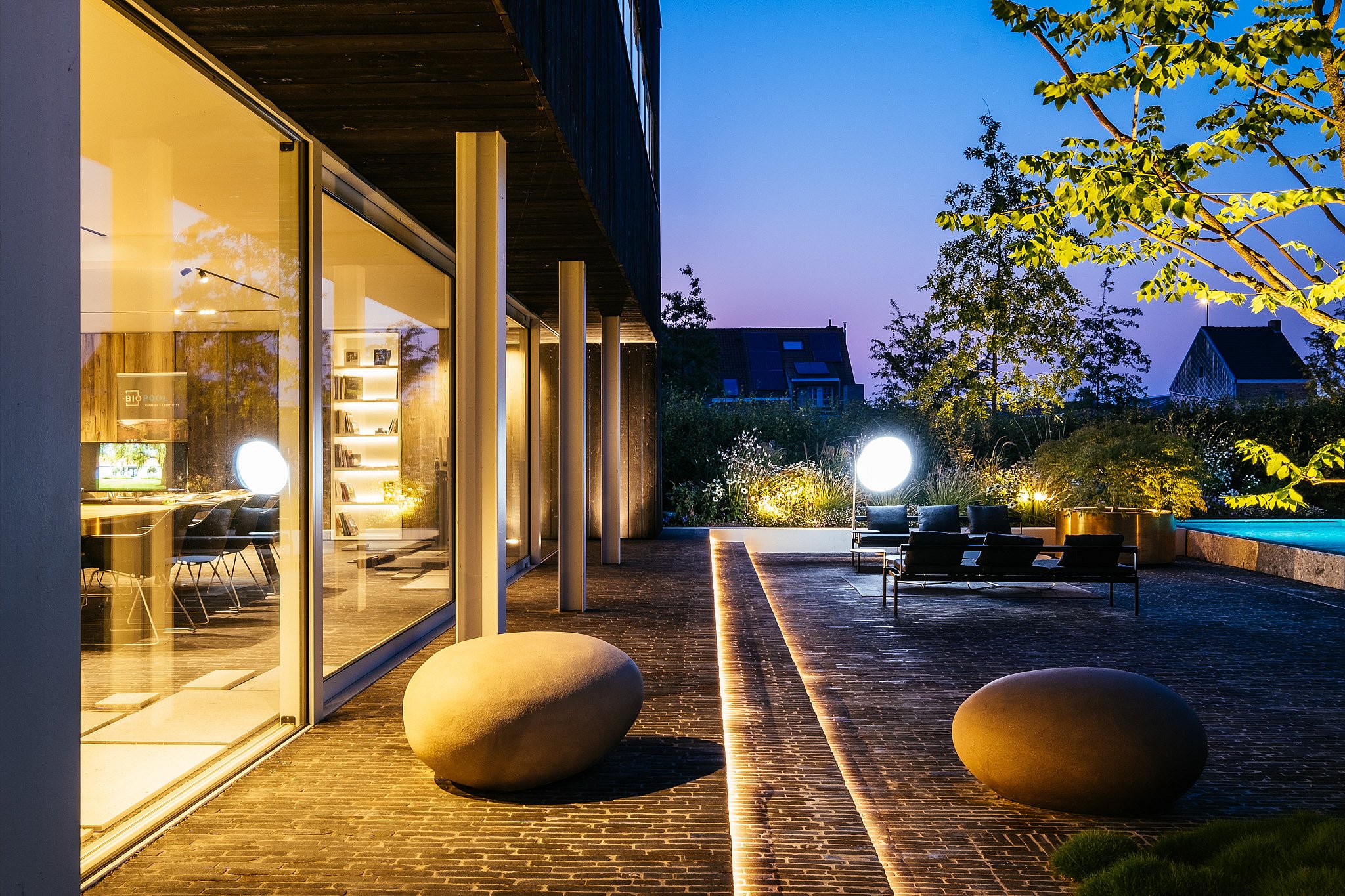 In der Abenddämmerung leuchtet die Terrasse des modernen Büros mit warmem Licht, während Sol+Luna-Loungesitze und runde beleuchtete Lampen eine entspannte Umgebung neben dem Pool schaffen.