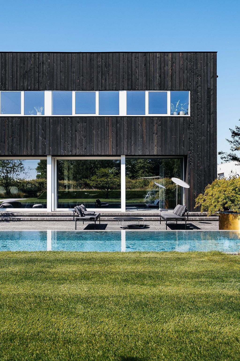 Ein dunkles, mit Holz verkleidetes Bürogebäude bietet Blick auf einen Pool. Auf der Terrasse vor der Glasfassade sind zwei Sol+Luna-Loungesitze und niedrige Tische angeordnet.