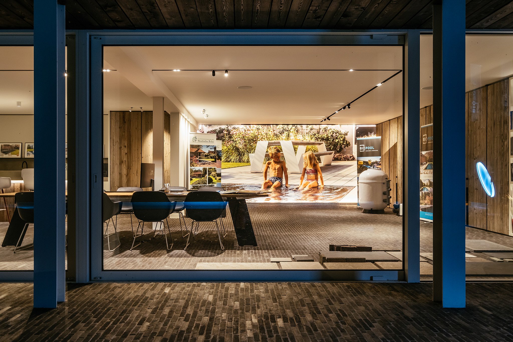 Blick durch ein großes Fenster in den warm beleuchteten Monbaliu-Showroom mit einem Besprechungstisch und Stühlen im Inneren und einer Gartenplanungsausstellung auf der Rückseite