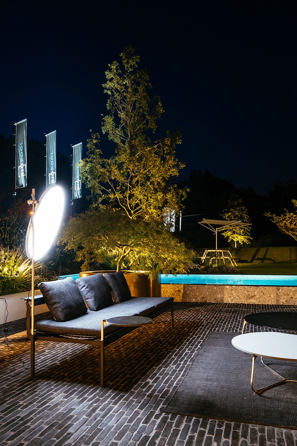 Nahaufnahme eines dunkelgrauen Sol+Luna-Sofas auf der Terrasse bei Nacht, beleuchtet von einer leuchtenden runden Lampe, mit dem Pool und einem Virus-Tisch mit Akazienschirm im Hintergrund.