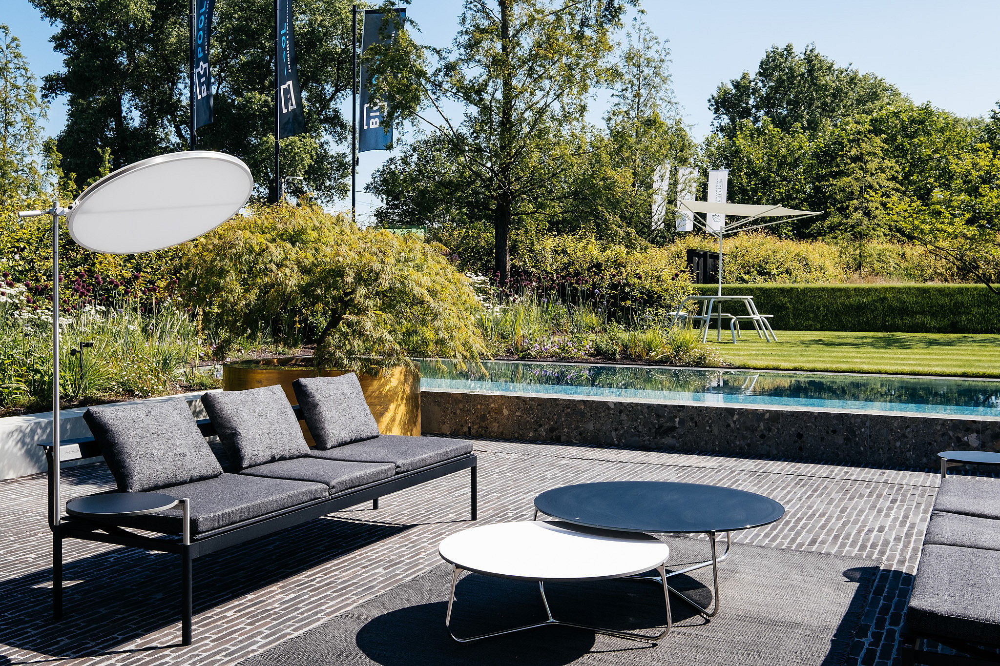 Donkergrijze Sol + Luna lounge banken met ronde bijzettafels zitten op een verhard terras naast een lang zwembad, met een witte schijfvormige lamp en een Virus tafel met Acacia schaduw zichtbaar in de aangelegde tuin daarbuiten.