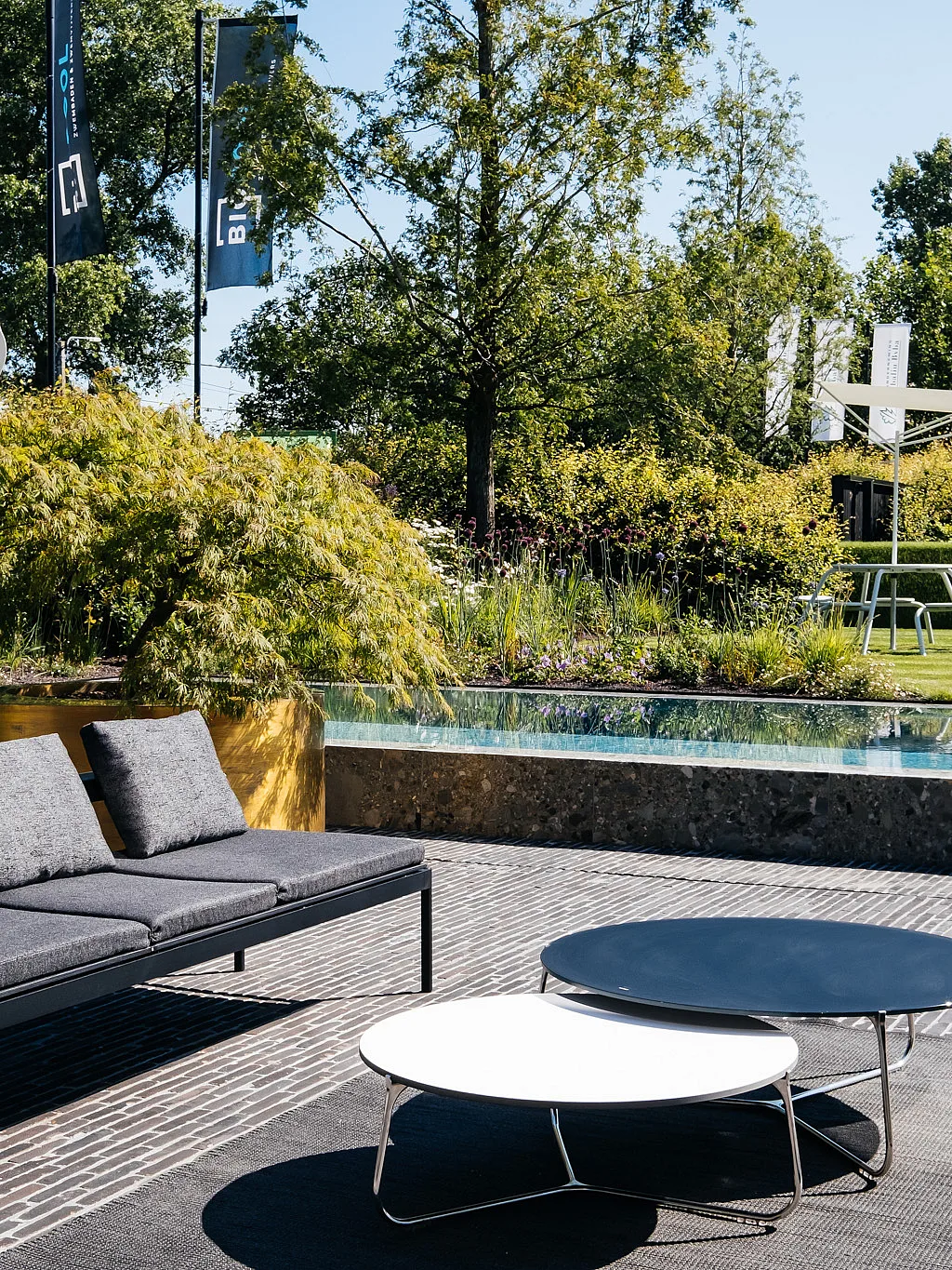 Les canapés lounge gris foncé Sol+Luna avec des tables rondes sont installés sur une terrasse pavée au bord d’une longue piscine, avec une lampe blanche en forme de disque et une table Virus avec un abat-jour en acacia visible dans le jardin paysager au-delà.