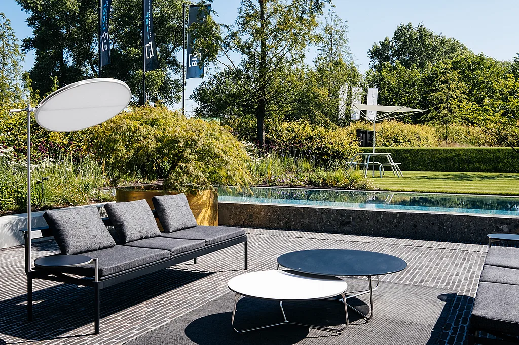Dark grey Sol+Luna lounge sofas with round side tables sit on a paved terrace beside a long pool, with a white disc-shaped lamp and a Virus table with Acacia shade visible in the landscaped garden beyond.