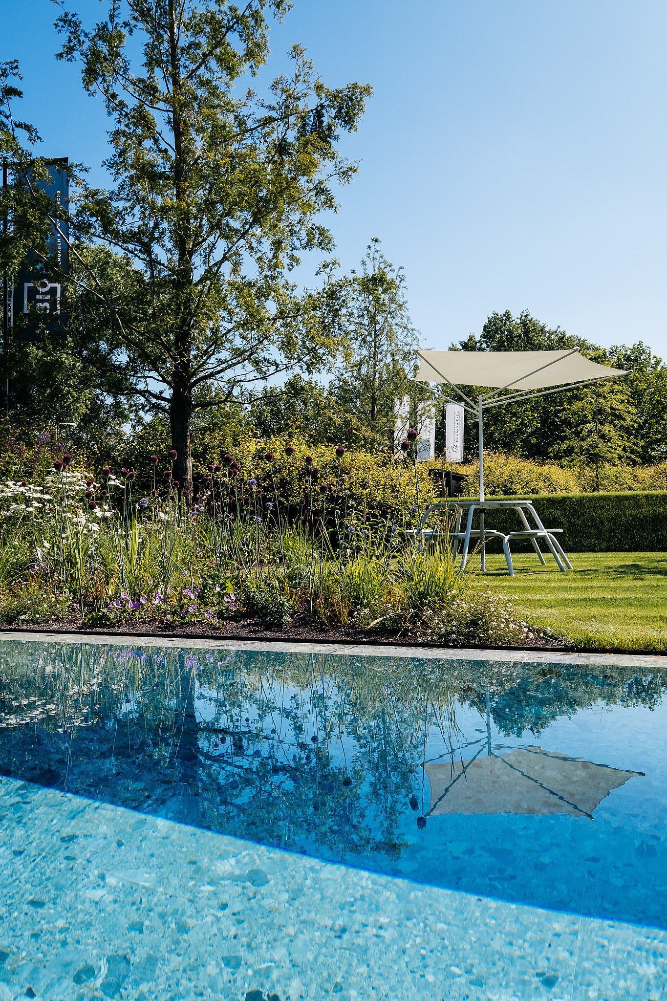 Ein blasser Virus-Picknicktisch mit integrierten Hockern steht auf dem Rasen unter einem passenden Akazienton, der sich im blauen Pool neben blühenden Rändern widerspiegelt.