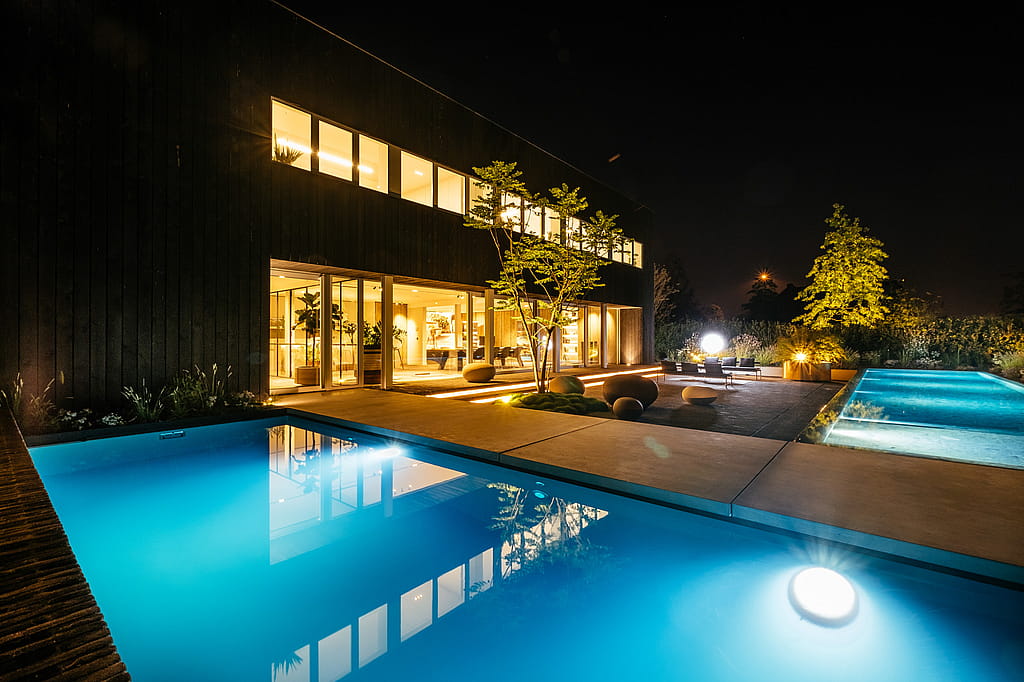 Nachtblick über den leuchtenden Pool zur Terrasse, wo Sol+Luna-Loungesitze unter beleuchteten Bäumen und einer hellen runden Lampe sitzen.