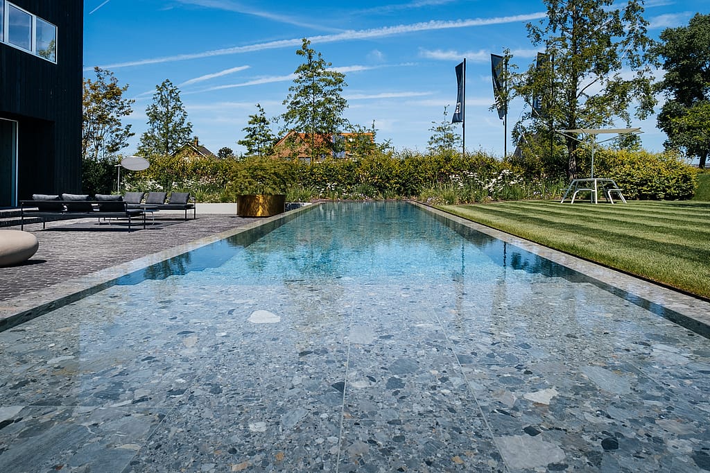 Weite Aussicht auf einen modernen Garten mit einem langen reflektierenden Pool, Sol+Luna-Lounge-Sitzen auf der Terrasse und einem hellen Virus-Tisch mit Akazienschatten auf dem gestreiften Rasen.