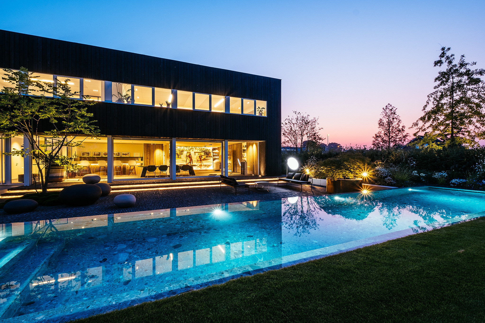 Abendlicher Blick auf das dunkel gekleidete Büro und den langen beleuchteten Pool, mit Sol+Luna-Sitzplätzen auf der Terrasse vor einem rosa-blauen Sonnenuntergangshimmel.