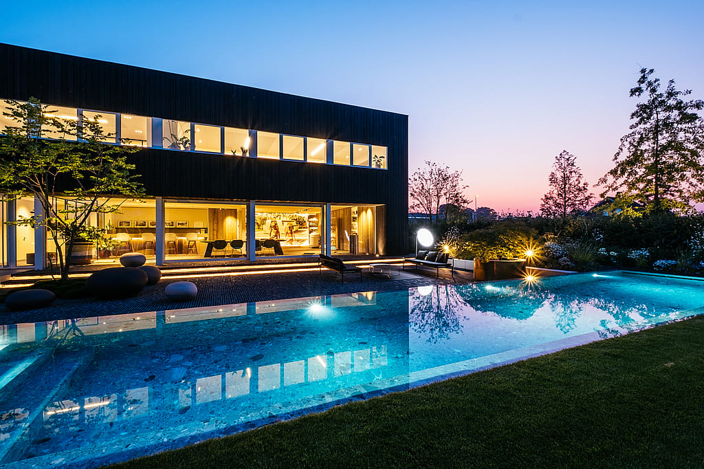 Abendlicher Blick auf das dunkel gekleidete Büro und den langen beleuchteten Pool, mit Sol+Luna-Sitzplätzen auf der Terrasse vor einem rosa-blauen Sonnenuntergangshimmel.
