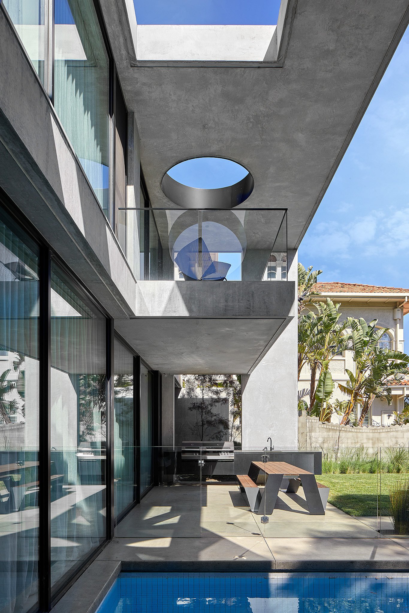 A modern concrete villa with a long blue pool and a Hopper picnic table on the terrace, framed by large sliding glass doors and round skylight openings.