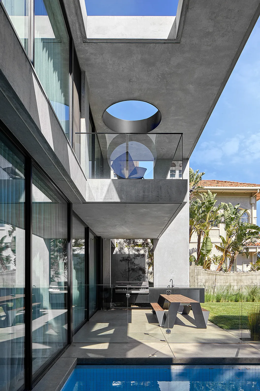 A modern concrete villa with a long blue pool and a Hopper picnic table on the terrace, framed by large sliding glass doors and round skylight openings.