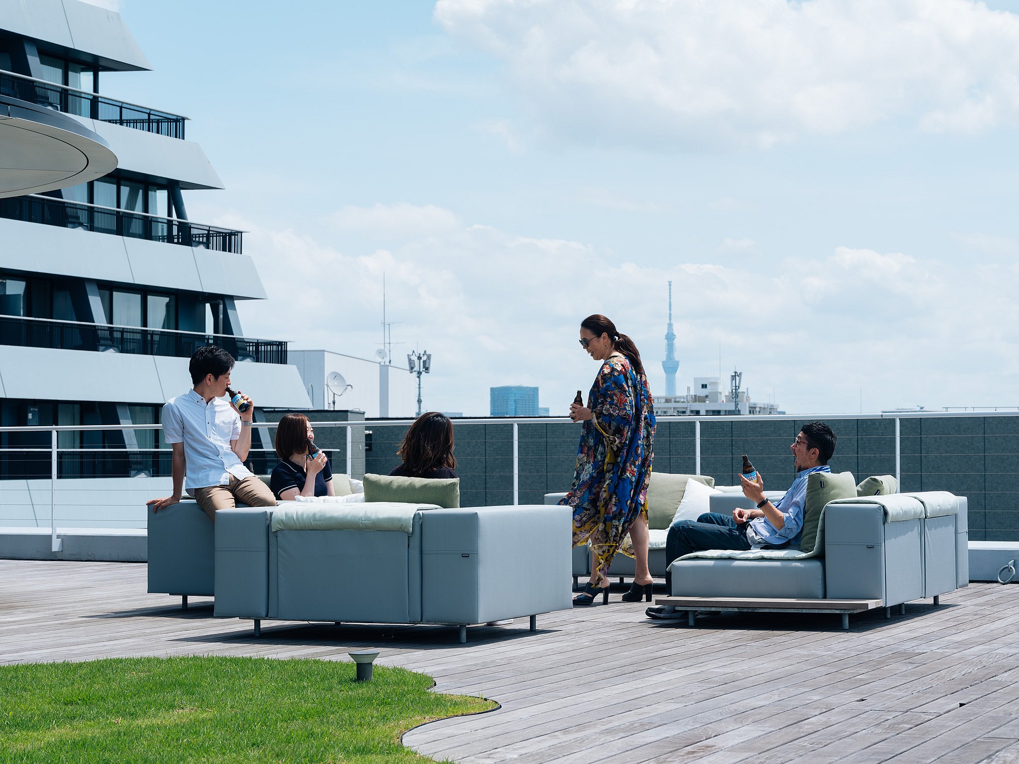 Cinq membres du personnel de l’ambassade se détendent avec des boissons en bouteille sur des canapés gris pâle Walrus sur une terrasse sur le toit, avec la ligne d’horizon de Tokyo et Tokyo Skytree visibles à l’arrière-plan.