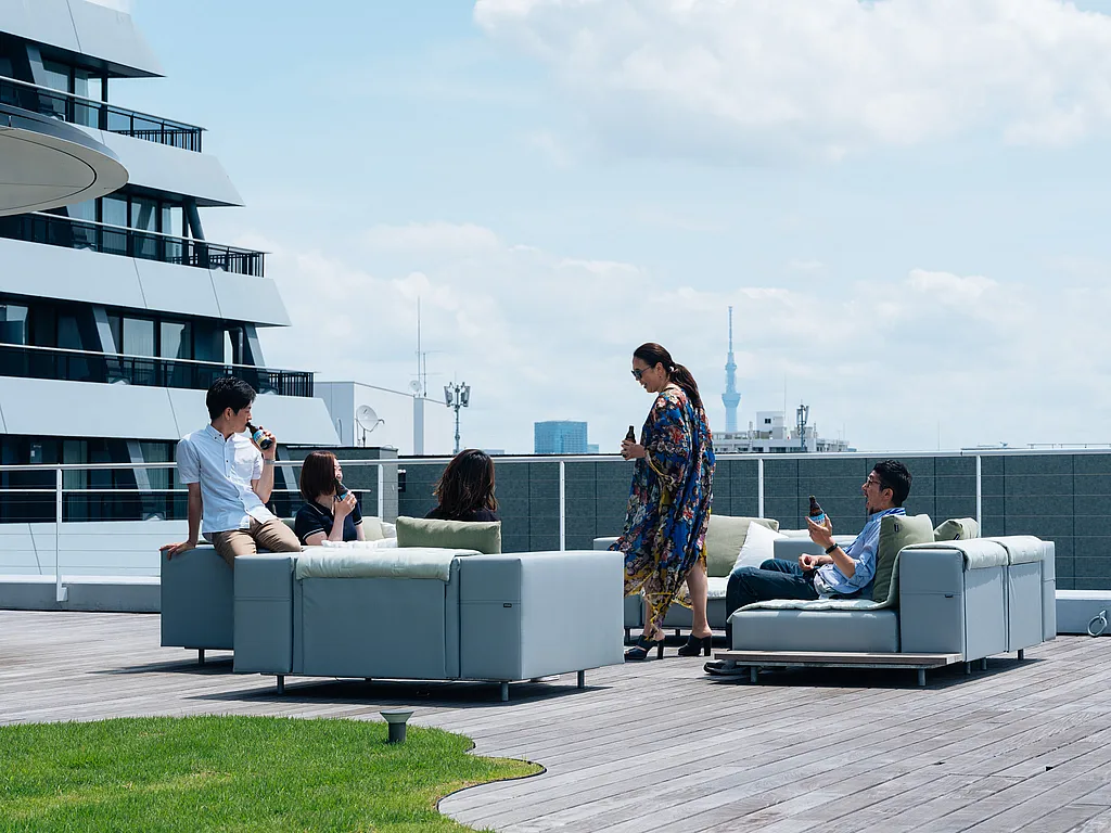 Cinq membres du personnel de l’ambassade se détendent avec des boissons en bouteille sur des canapés gris pâle Walrus sur une terrasse sur le toit, avec la ligne d’horizon de Tokyo et Tokyo Skytree visibles à l’arrière-plan.