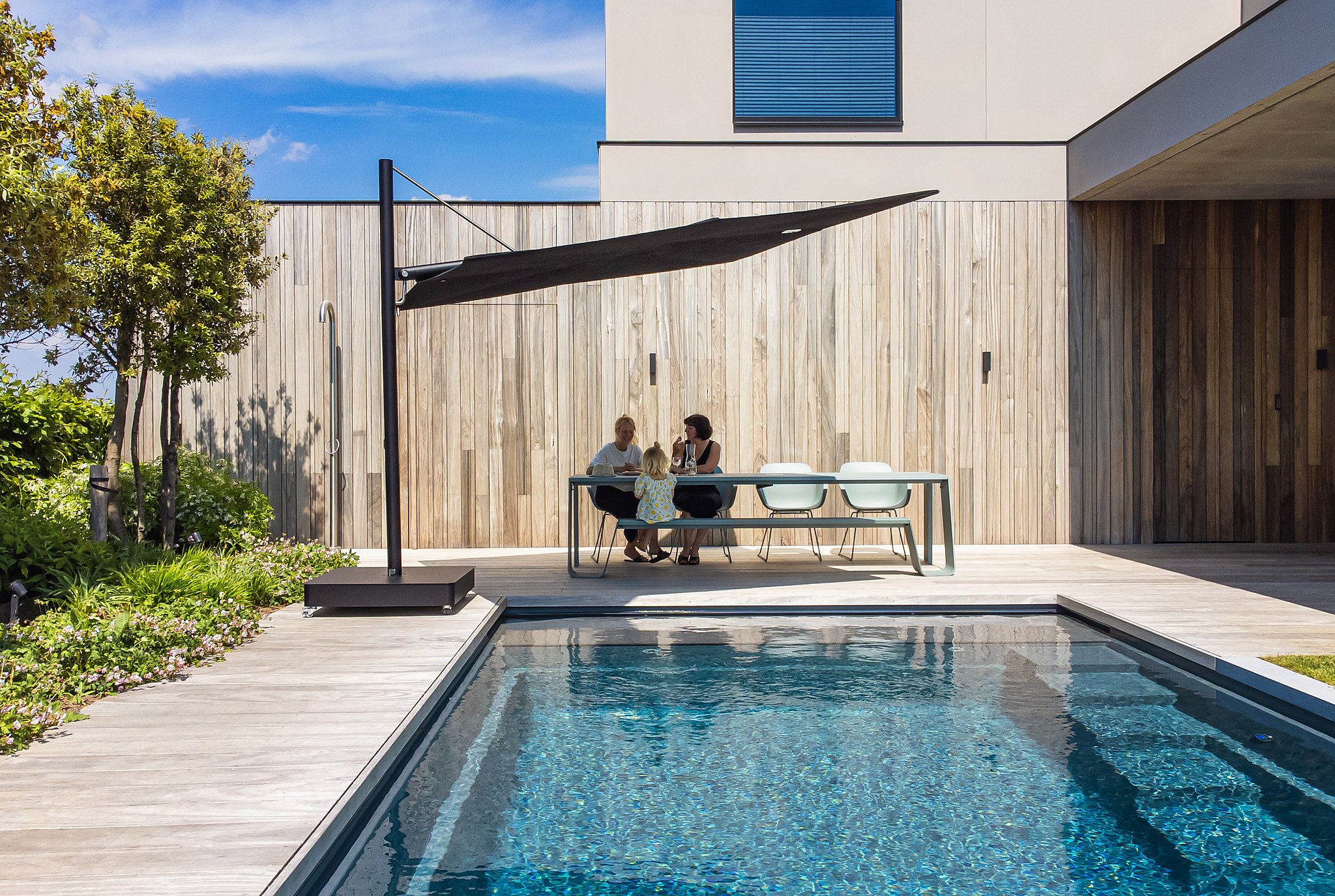A black Sensu sunshade shades a pale green Hopper combo table beside a narrow pool, where two women and a child sit on a sunlit terrace beside a modern house.