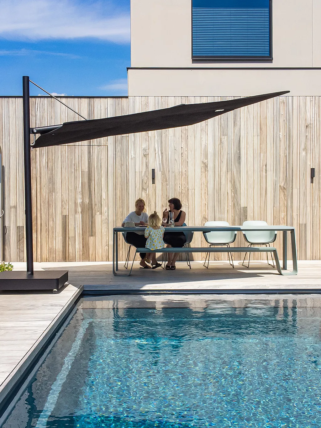 Een zwarte Sensu-zonnekap kleurt een lichtgroene Hopper-combinatietafel naast een smal zwembad, waar twee vrouwen en een kind op een zonovergoten terras naast een modern huis zitten.
