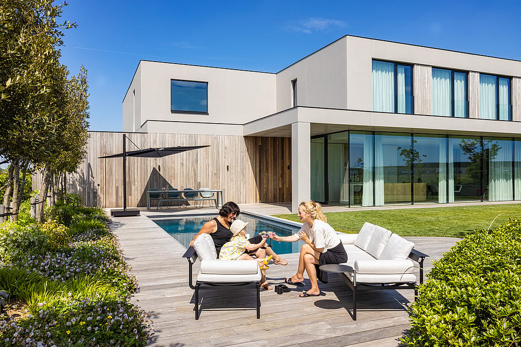 Twee vrouwen en een kind ontspannen op witte Sol + Luna loungestoelen naast het zwembad, met de Sensu-schaduwrijke eethoek en moderne villa erachter.