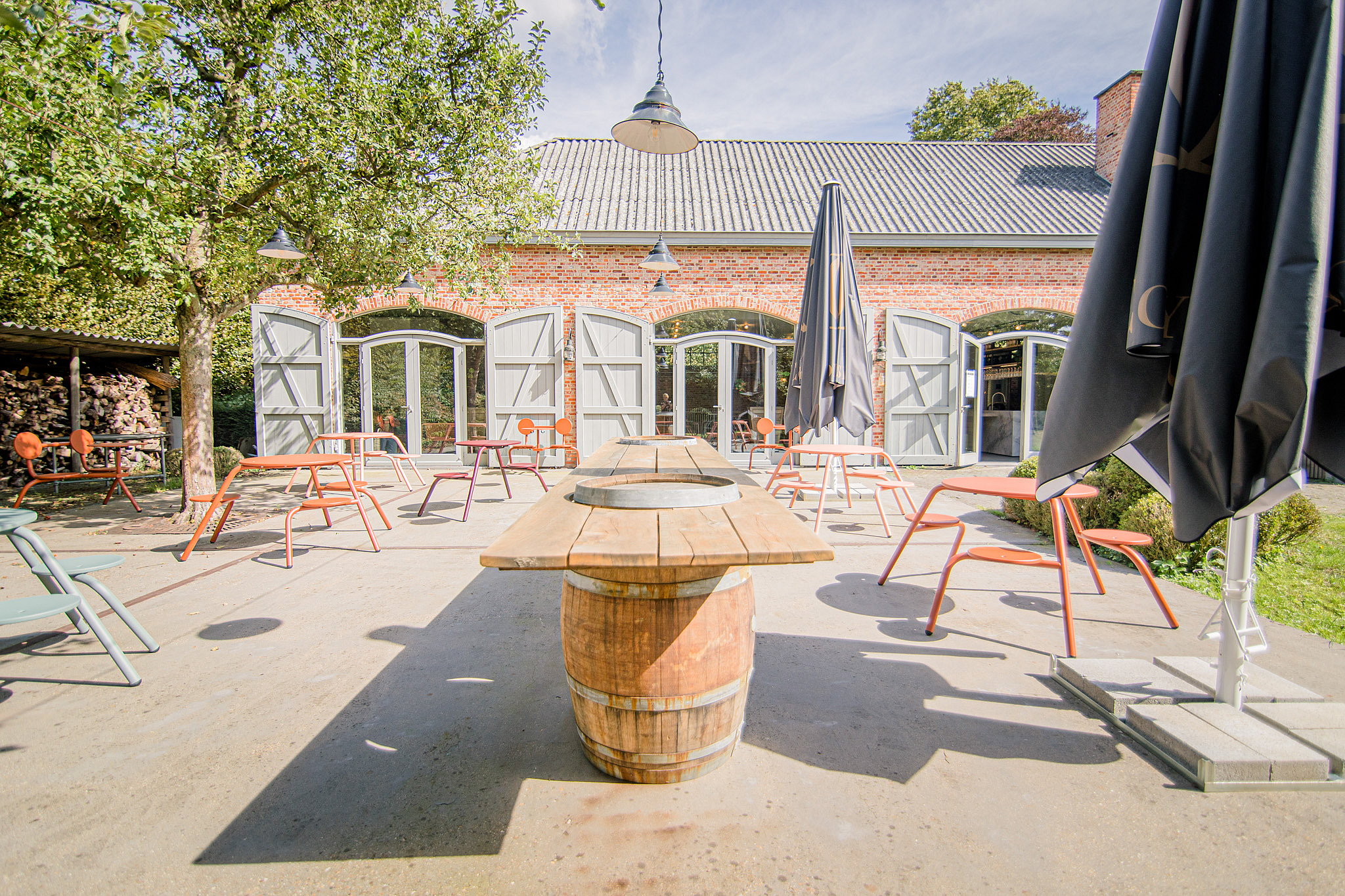 Verschillende kleurrijke cafévirustafels zijn verspreid onder volwassen bomen op de binnenplaats, met wijnflessen en glazen op de achtergrond.
