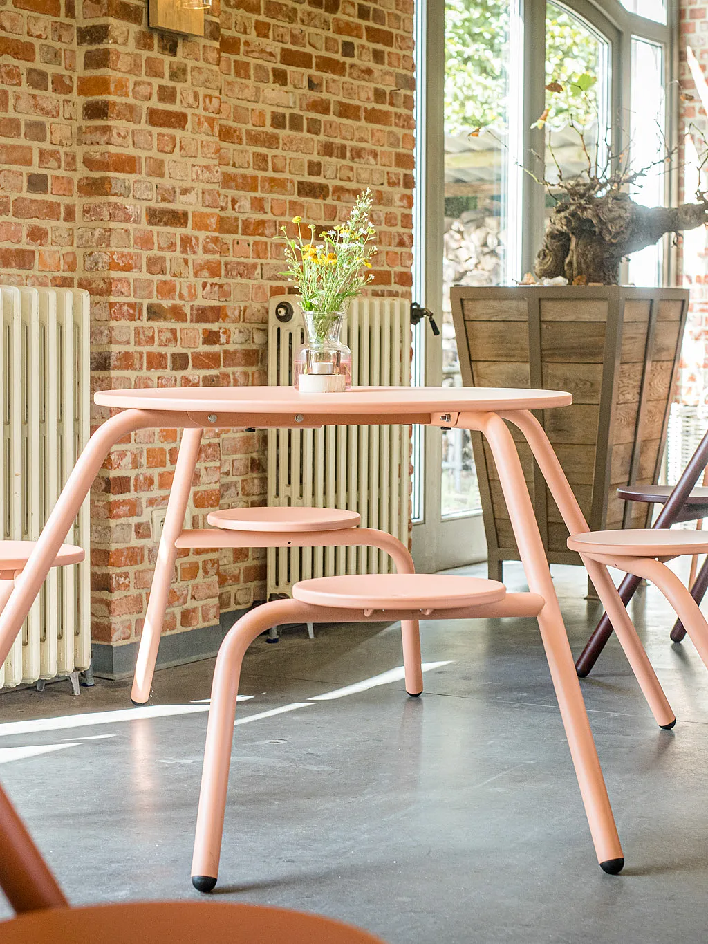 Een koraalroze café-tafel met geïntegreerde ronde stoelen staat in een zonovergoten bakstenen proefruimte, met kleurrijkere stoelen en planken op de achtergrond.