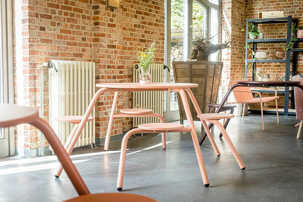 A coral-pink café table with integrated round seats stands in a sunlit brick tasting room, with more colorful seating and shelving in the background.