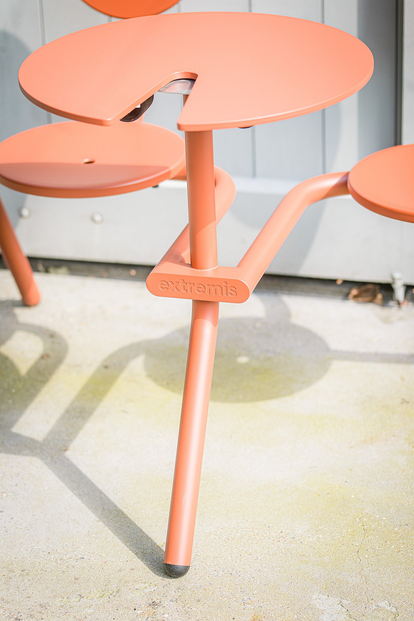 Close-up van een koraalkleurige ronde cafétafel met geïntegreerde stoelen, met de reliëfdruk Extremis-branding op het frame.