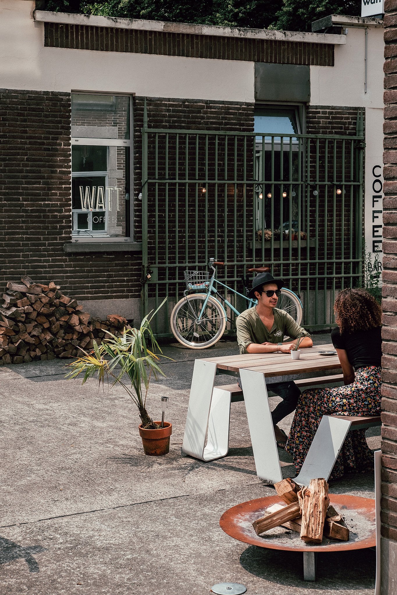 Deux personnes sont assises à discuter devant une table extérieure compacte en face du bar à café WATT, avec des piles de bois de chauffage, un vélo et un bol à feu à proximité.