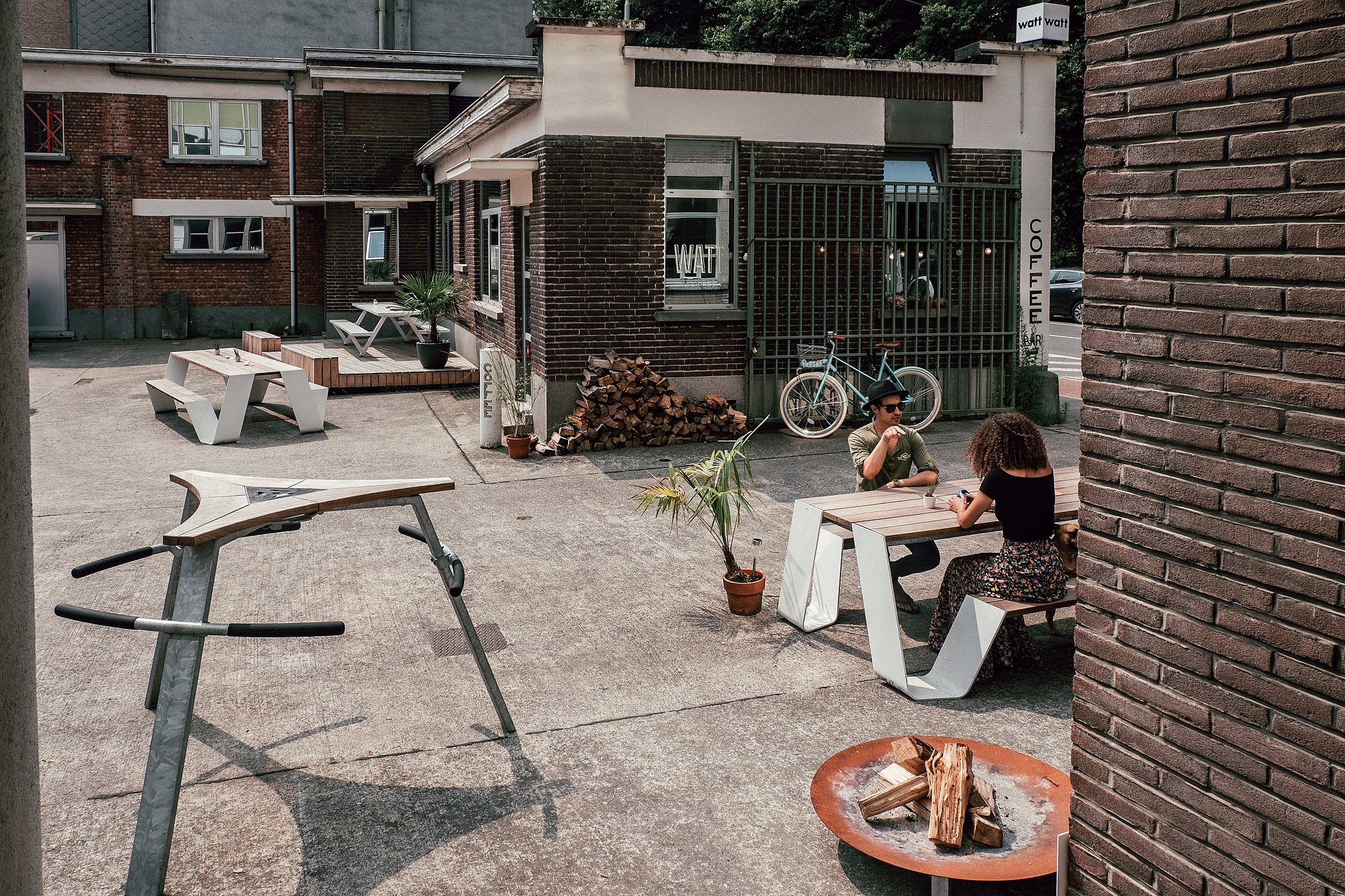 Breed uitzicht op de WATT-binnenplaats met witte buitentafels op een ruw betonnen oppervlak, waar twee mensen tegenover elkaar zitten aan een houten tafel naast een bakstenen koffiebar.