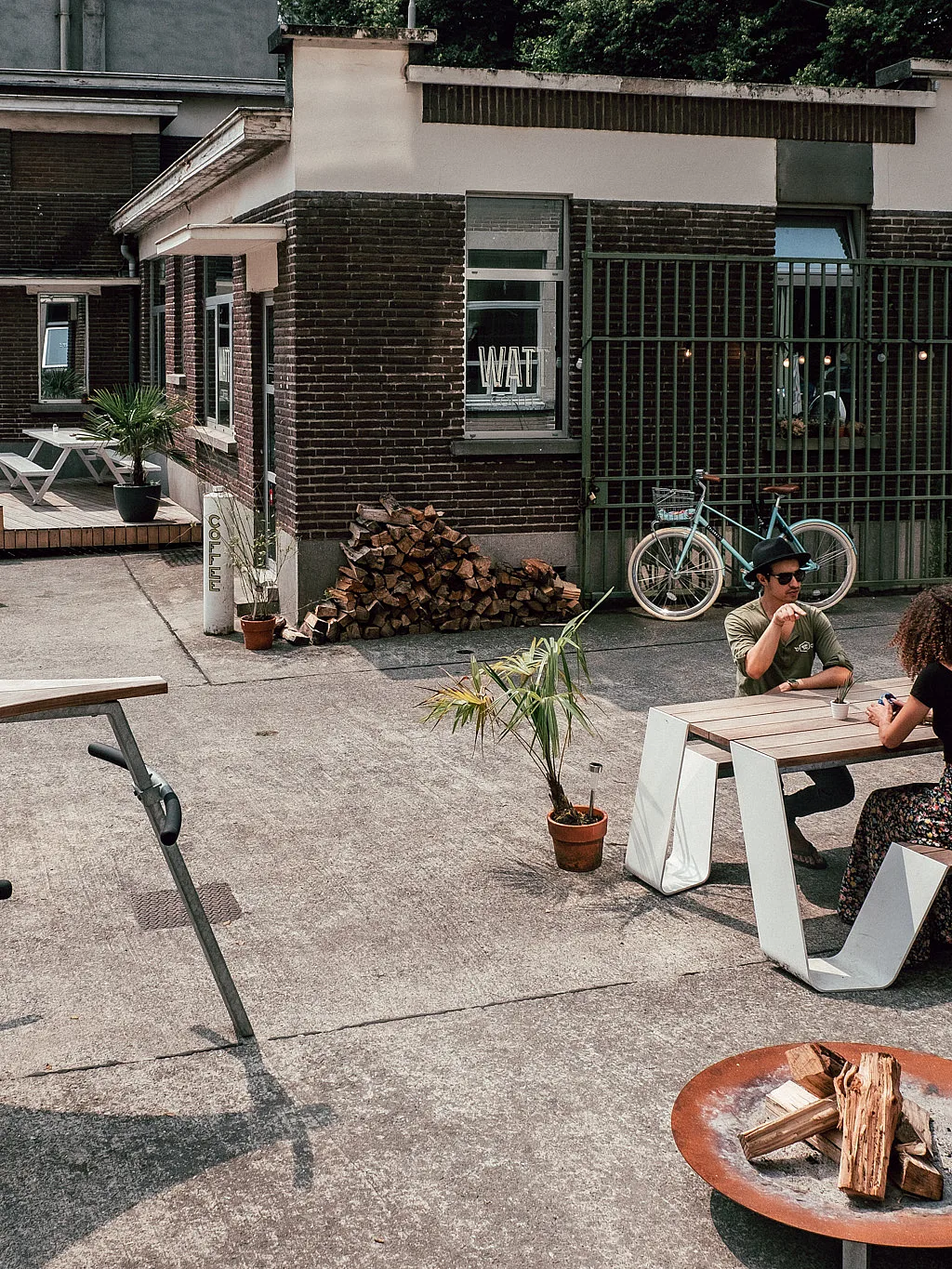 Breed uitzicht op de WATT-binnenplaats met witte buitentafels op een ruw betonnen oppervlak, waar twee mensen tegenover elkaar zitten aan een houten tafel naast een bakstenen koffiebar.