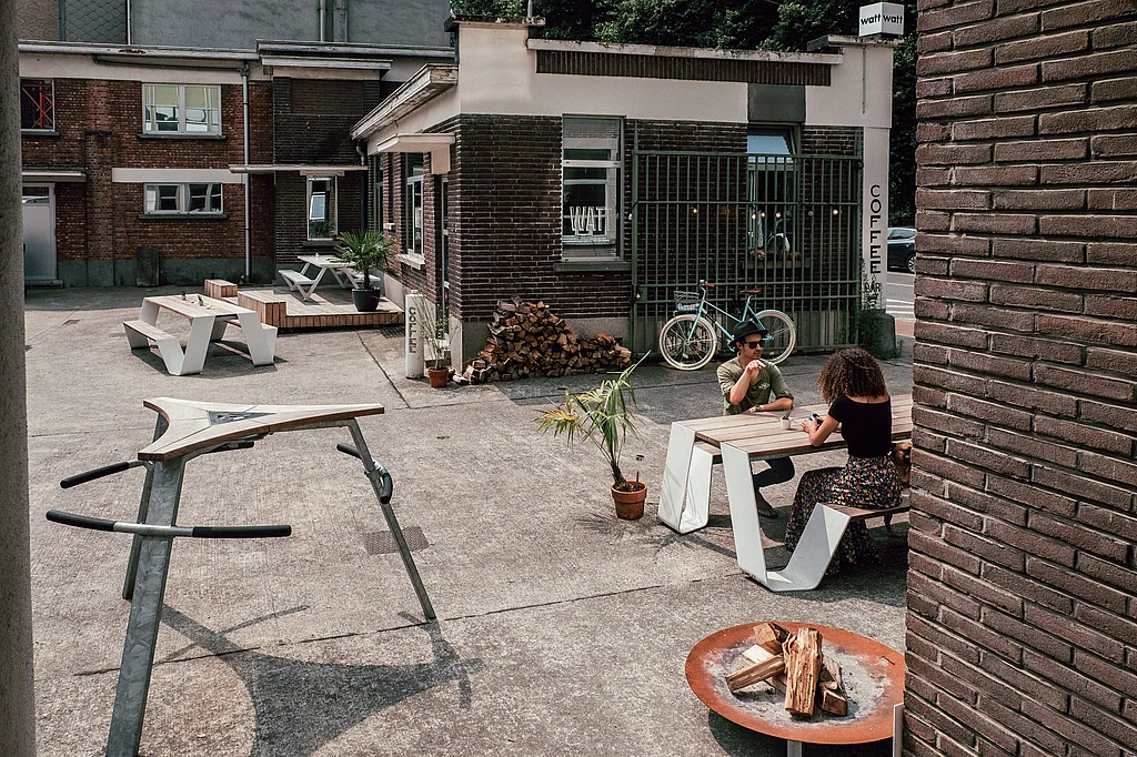 Breed uitzicht op de WATT-binnenplaats met witte buitentafels op een ruw betonnen oppervlak, waar twee mensen tegenover elkaar zitten aan een houten tafel naast een bakstenen koffiebar.