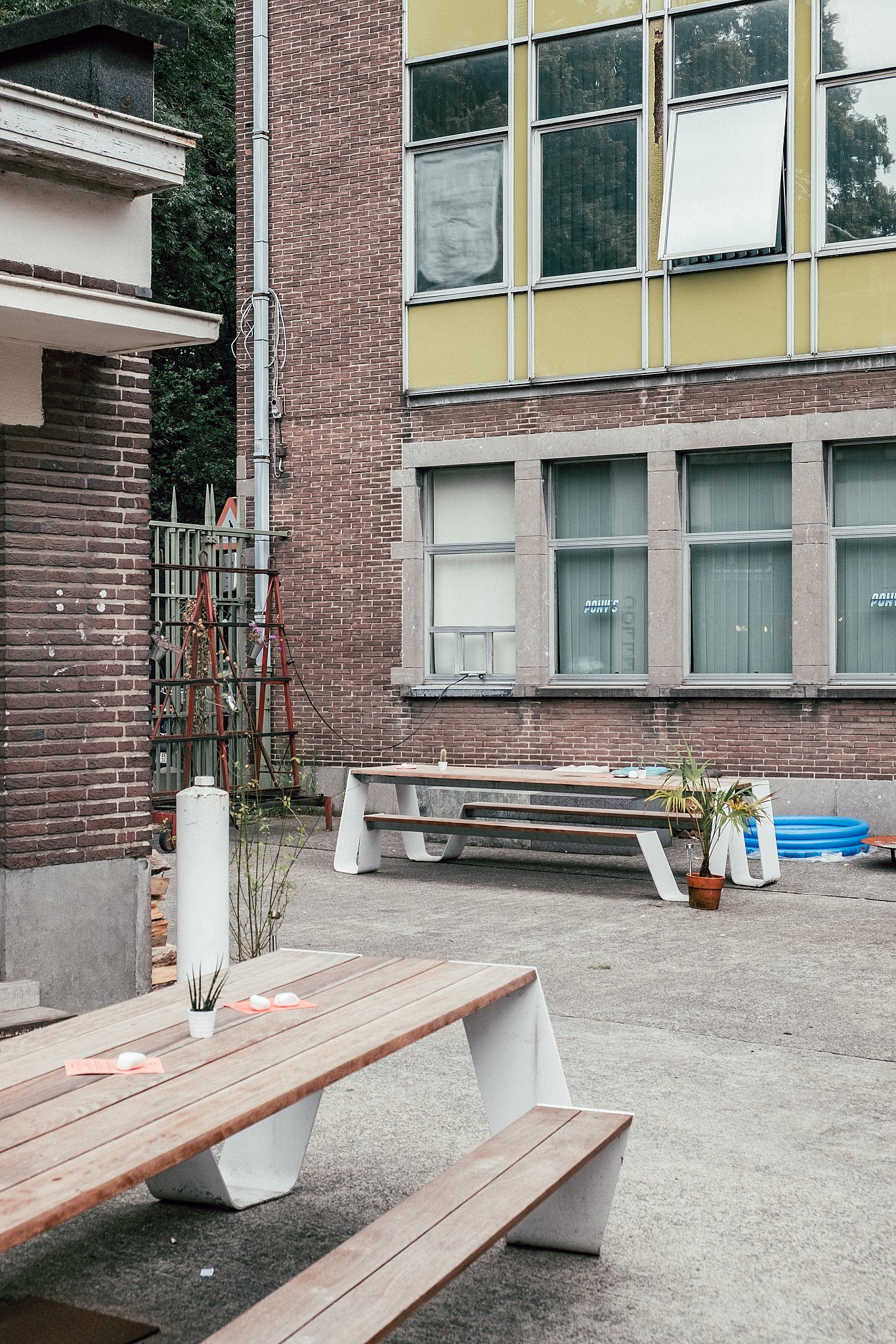 Deux tables trémies extérieures en bois se dressent sur la cour en béton à côté du bâtiment en briques, décorées de petites plantes et d’objets rose pâle sur les plateaux.