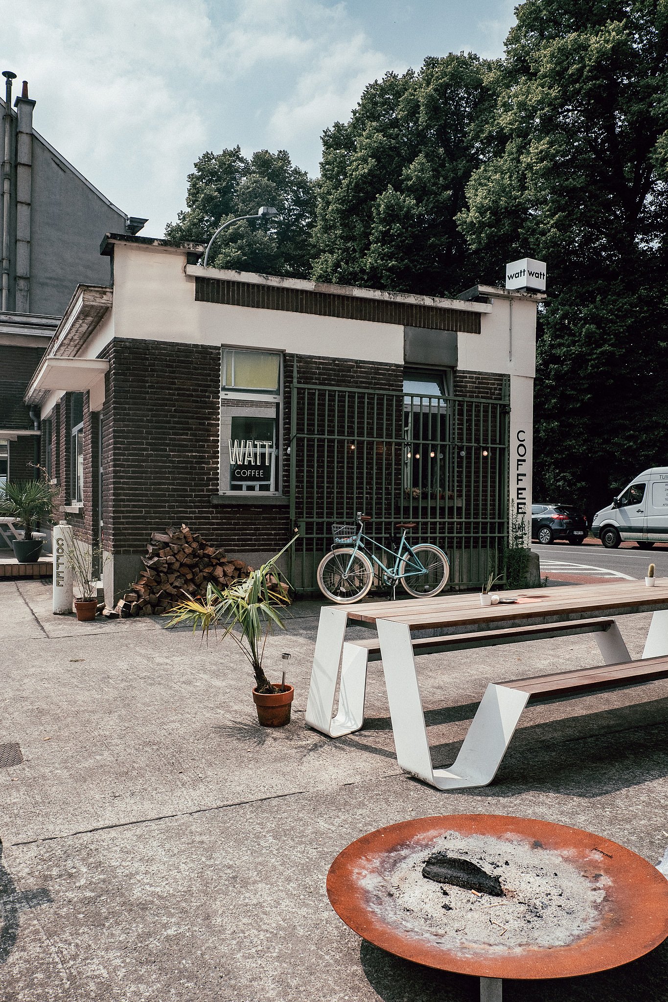 Une table et un banc en bois se dressent dans la cour devant le bar à café WATT, avec un vélo contre la façade en brique fermée et un bol à feu au premier plan.