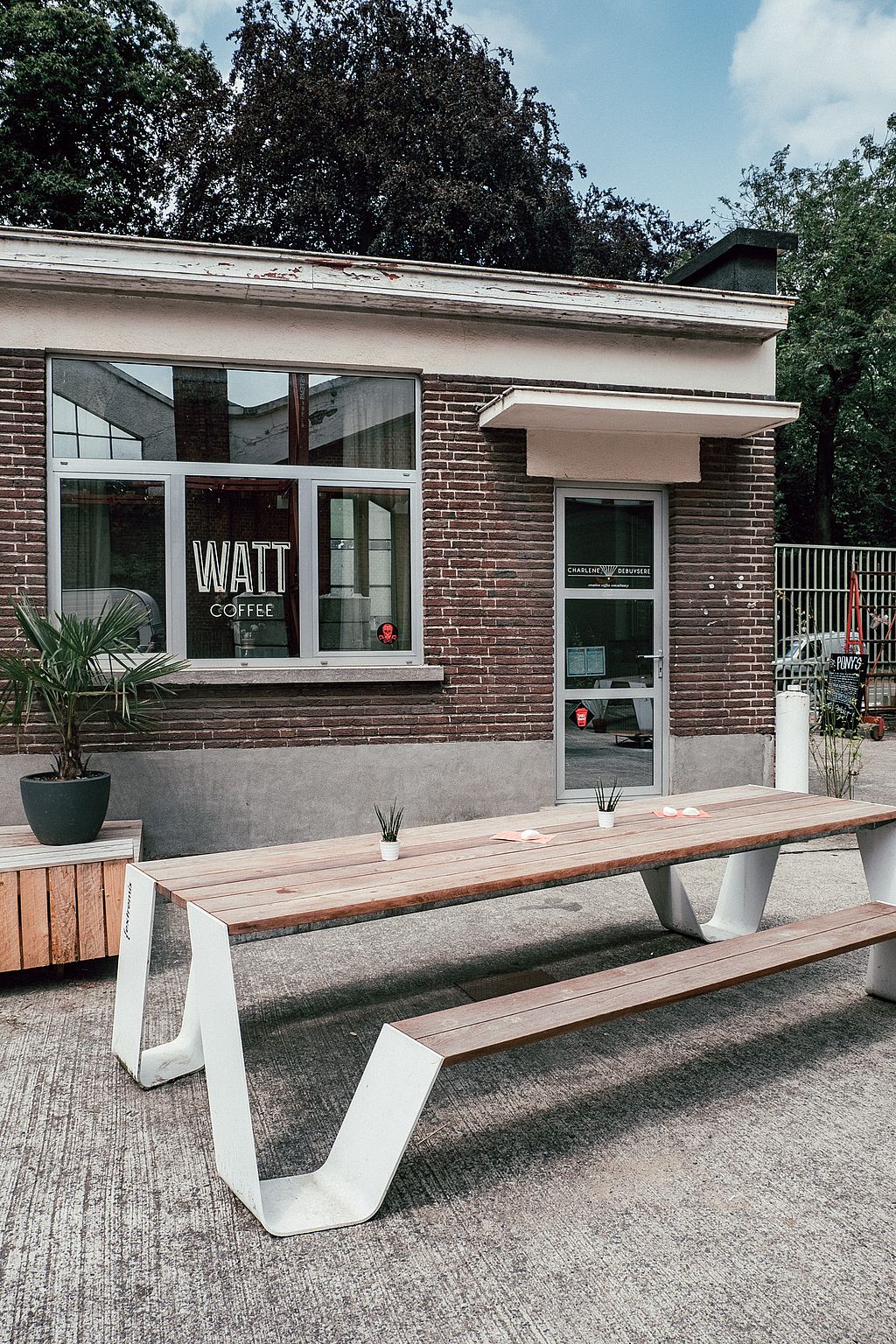 Vue rapprochée d’une longue table extérieure en bois avec banc assorti devant la fenêtre et l’entrée du bar à café WATT.