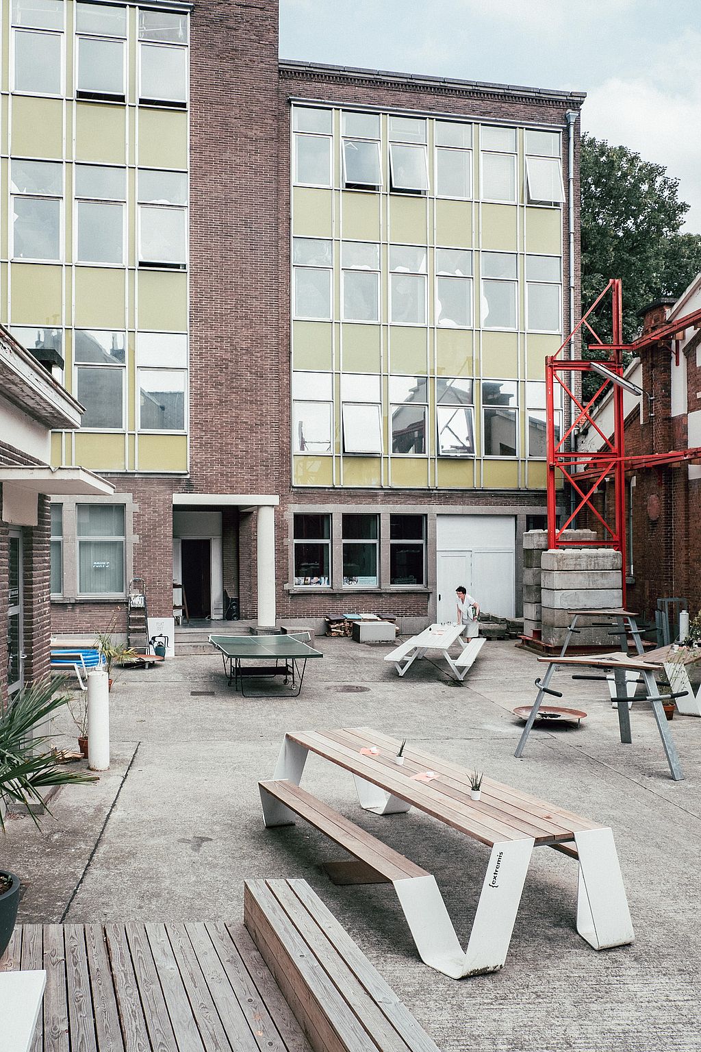 Plusieurs tables trémies extérieures blanches sont dispersées dans la cour industrielle du bâtiment WATT, avec une table de ping-pong et une haute façade en briques et verre à l’arrière-plan
