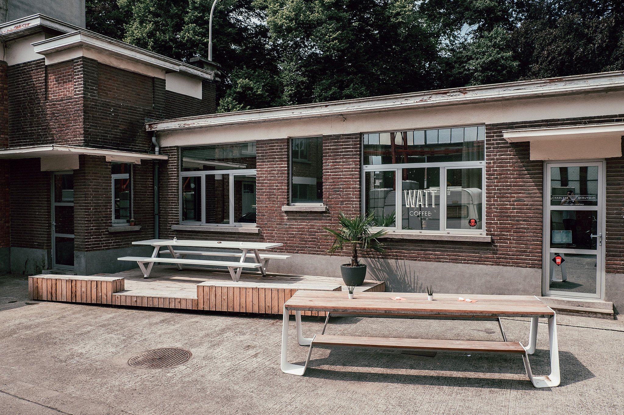 Une longue table trémie extérieure en bois se dresse sur la cour en béton devant le café WATT en brique, avec une table de pique-nique blanche sur une plateforme en bois derrière.