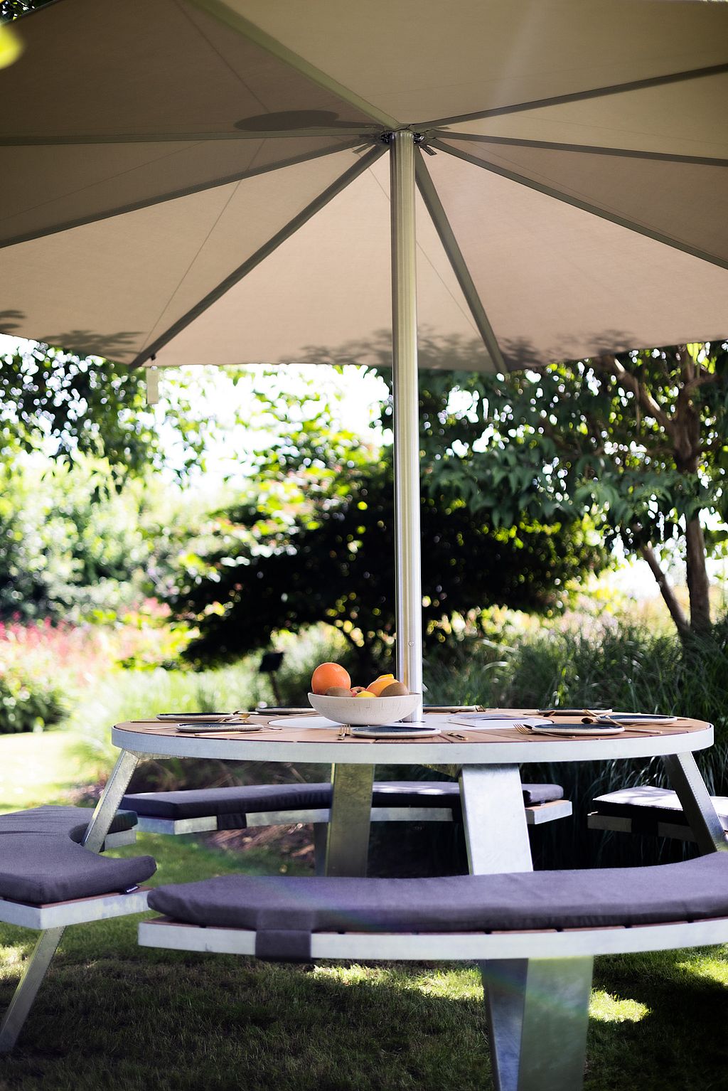 Close-up of a set Pantagruel table with curved benches under the wide canopy of an Inumbra shade, with a bowl of fruit at the centre.