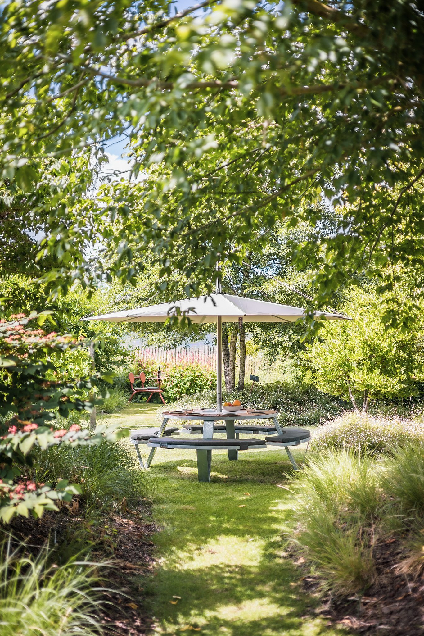 A Pantagruel picnic table beneath an Inumbra shade is seen at the end of a grassy path, surrounded by layered planting and overhanging branches.