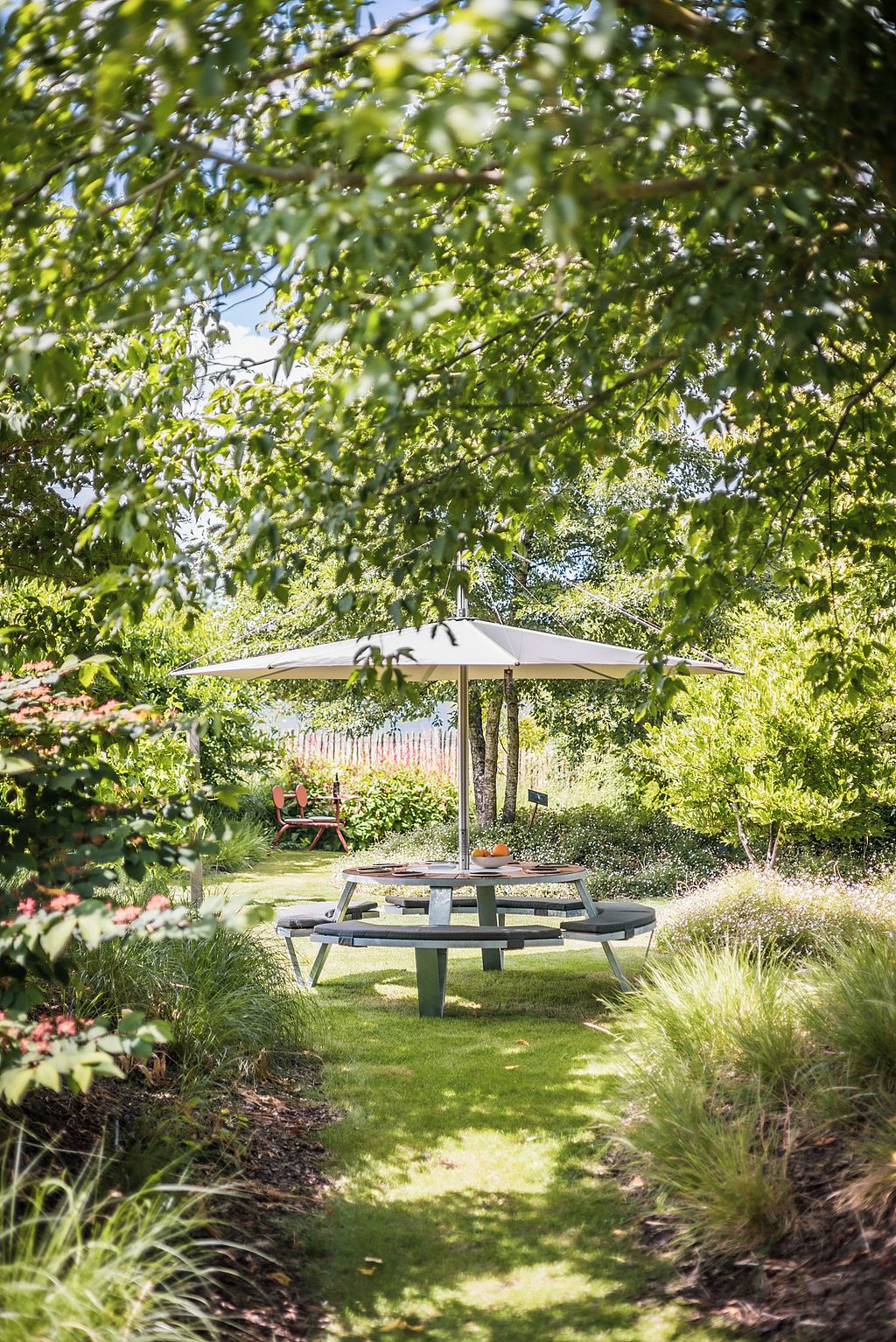 A Pantagruel picnic table beneath an Inumbra shade is seen at the end of a grassy path, surrounded by layered planting and overhanging branches.